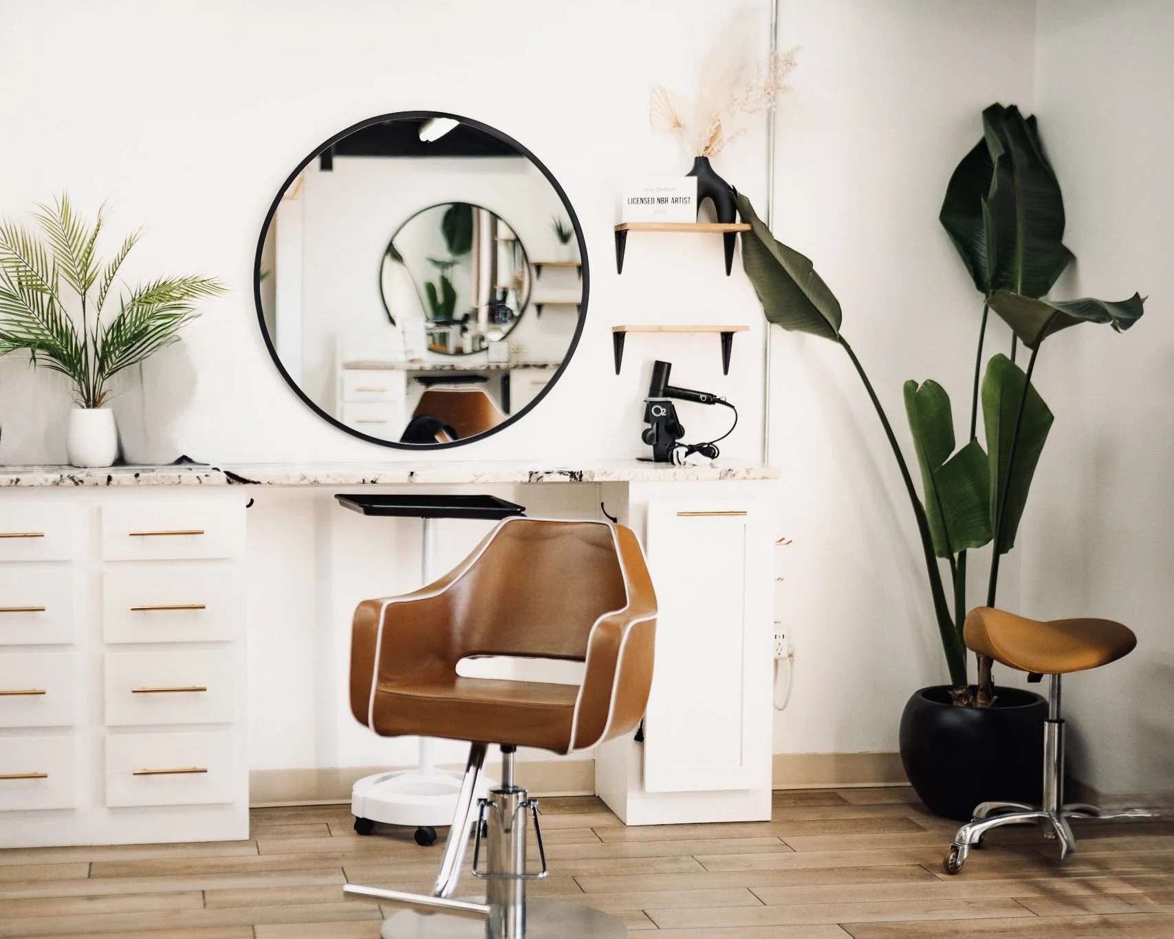 Modern hair salon with a brown stylist chair, large mirror, white cabinets, potted plants, and shelves with decorative items.