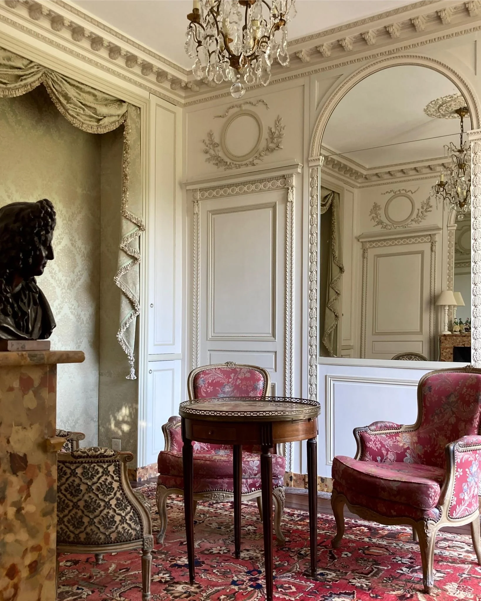 Intérieur d'une pièce élégante avec des meubles anciens, des miroirs, un lustre en cristal, une sculpture de bronze d'une femme, des rideaux brodés, des chaises rembourrées en tissu rose antique, un tapis orientale et des murs décorés en stuc ornés de moulures classiques.