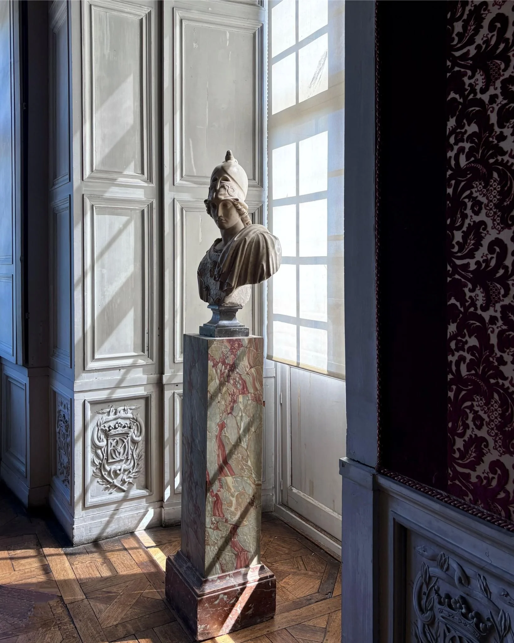 Une sculpture de buste de grec antique, placée sur un socle en marbre, dans une pièce avec des murs ornés et un sol en parquet.