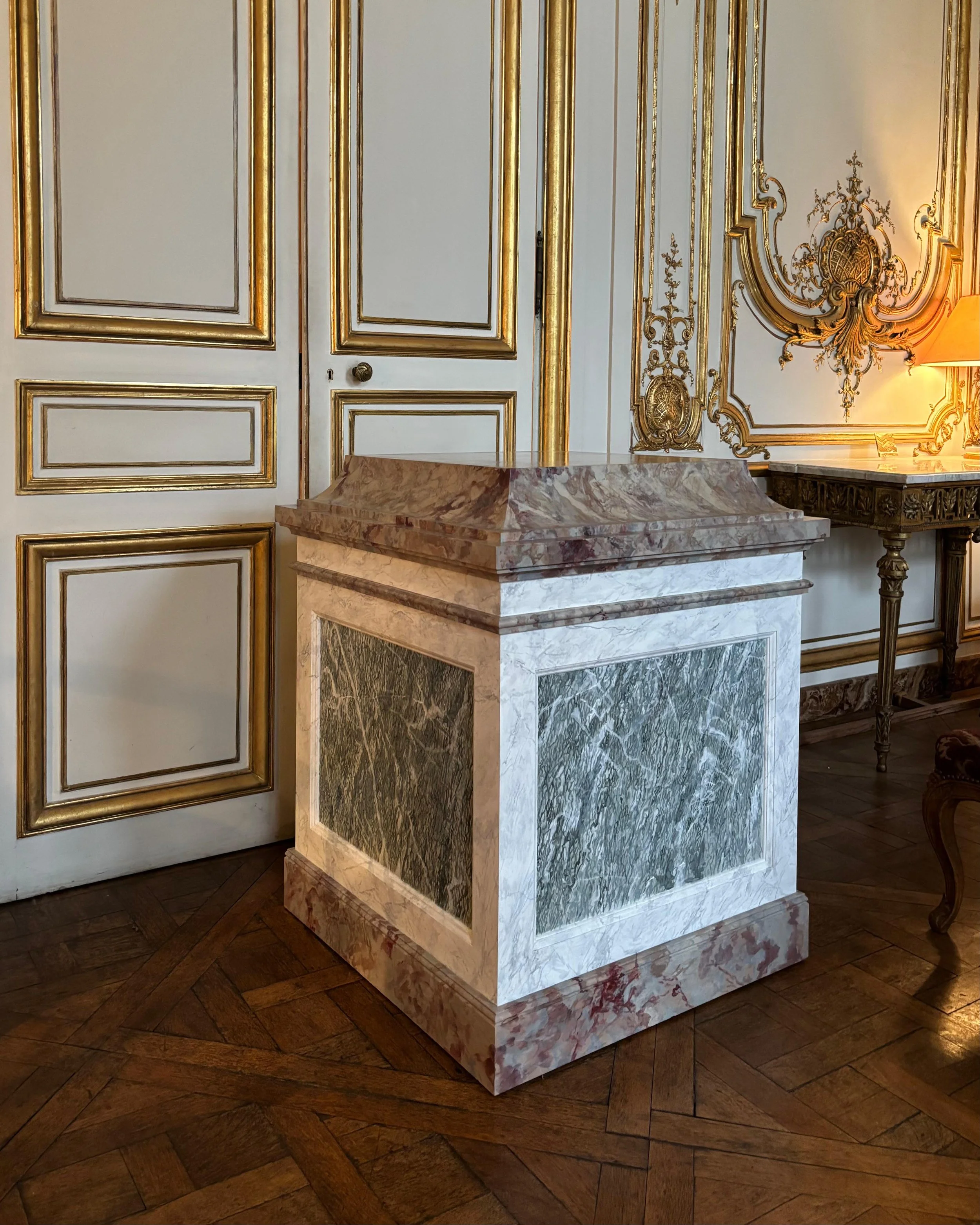 Socle en faux-marbre dans une pièce ornée de moulures dorées et de boiseries élégantes.