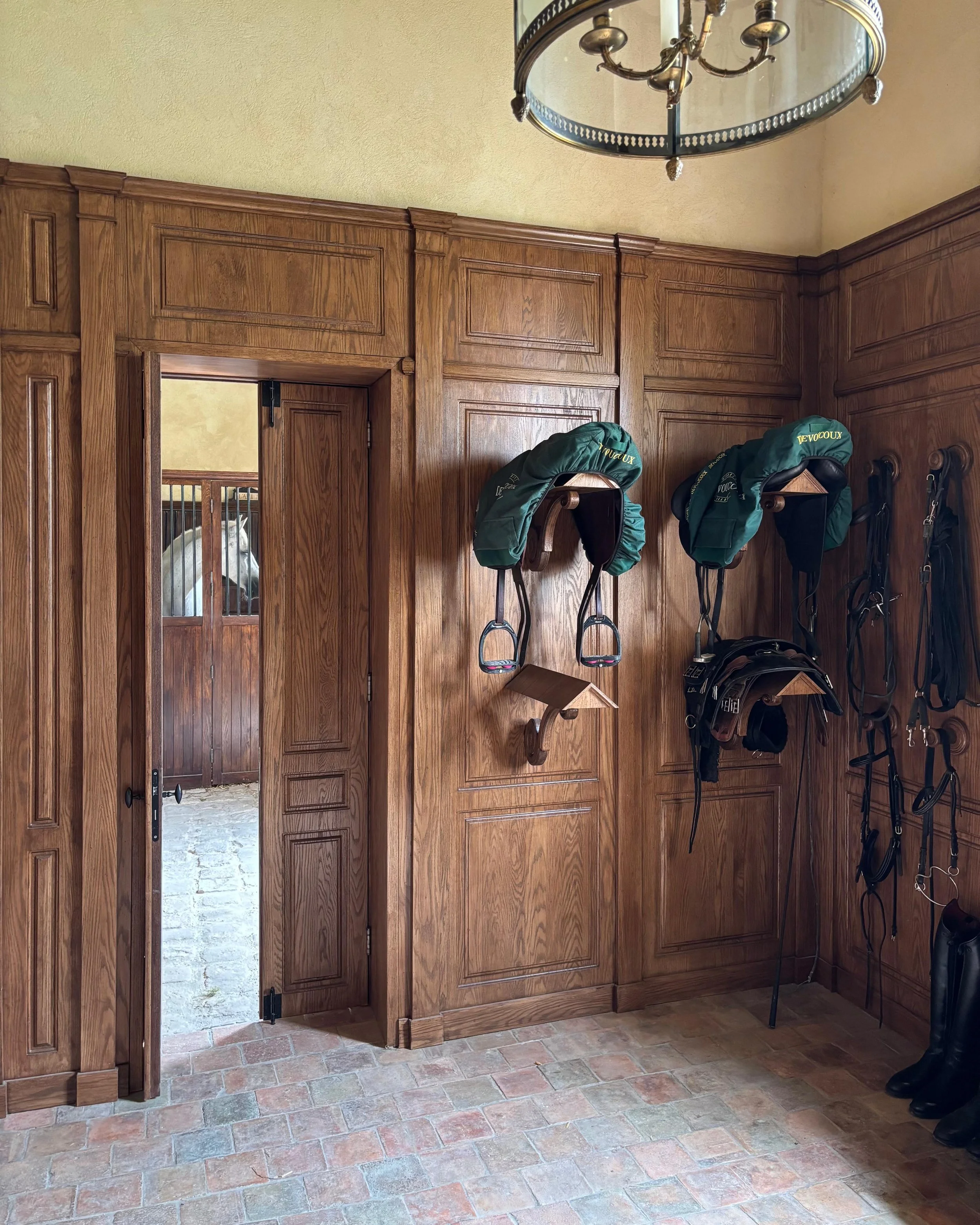 Une sellerie avec un mur en bois où sont suspendus des équipements d'équitation, dont des selles, des brides, des bottes, et un cheval blanc visible à travers une ouverture dans le mur.