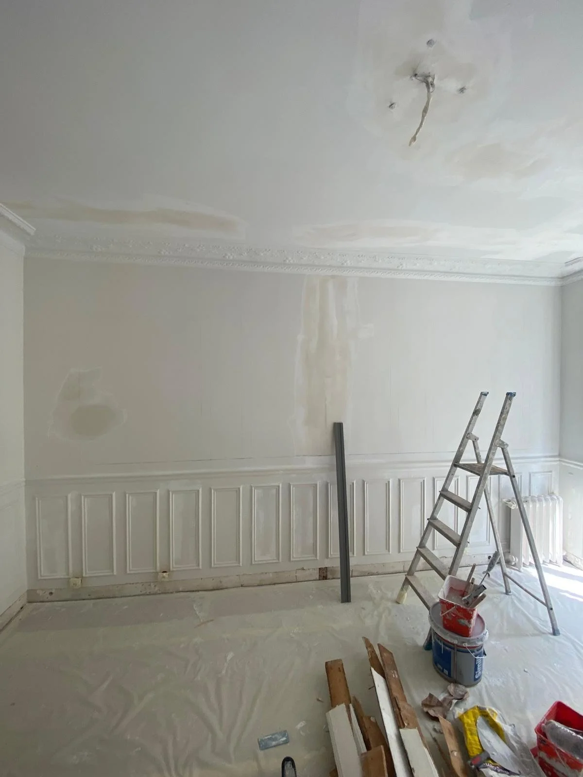 Une pièce en cours de rénovation avec un plafond et des murs en train d'être peints ou réparés, un escabeau en aluminium, des outils et des matériaux de peinture au sol.