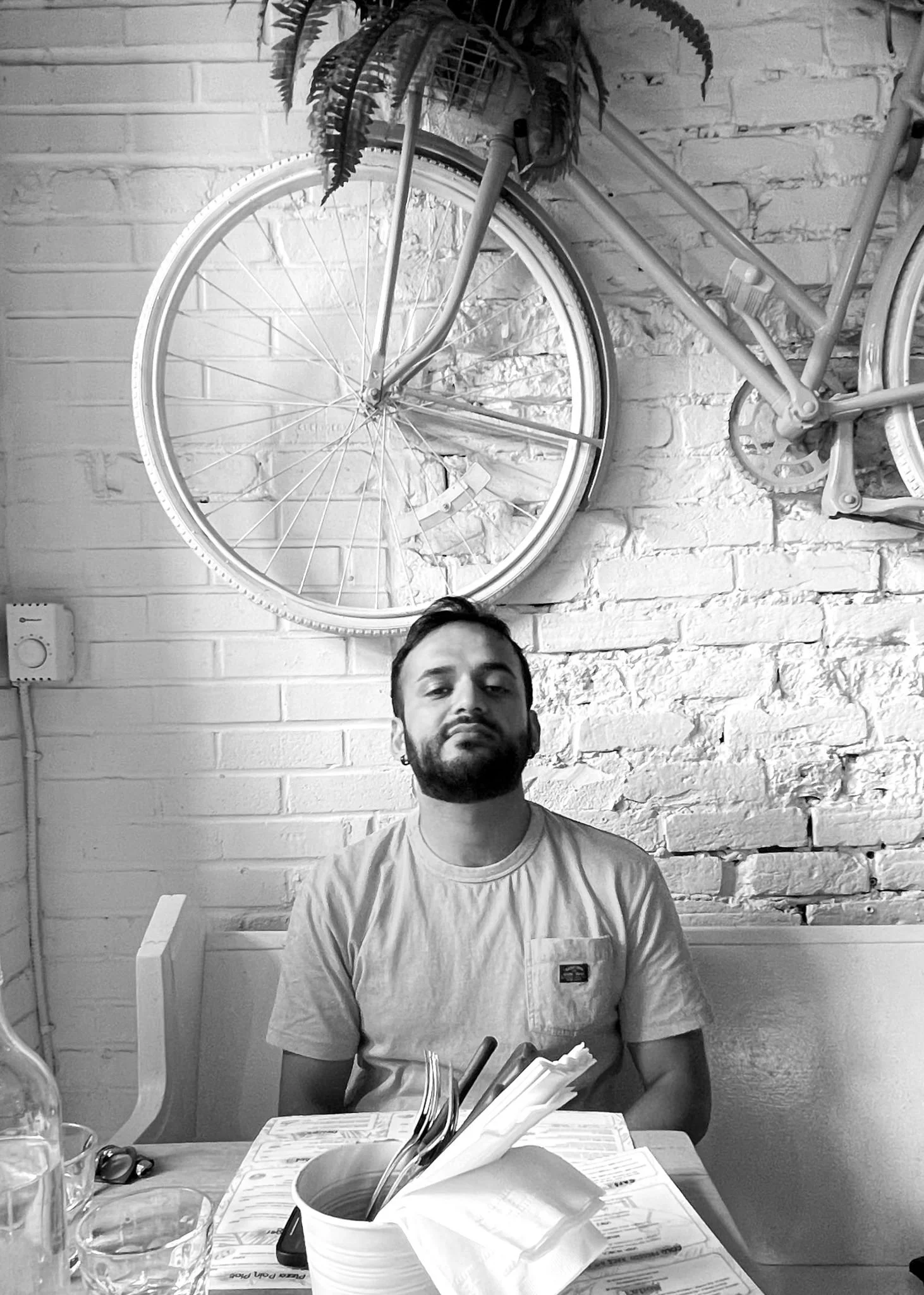 Saqib Malik est assis à une table dans un restaurant, avec un mur en briques blanches derrière lui, décoré d'un vélo fixé au mur et d'une plante en pot suspendue.