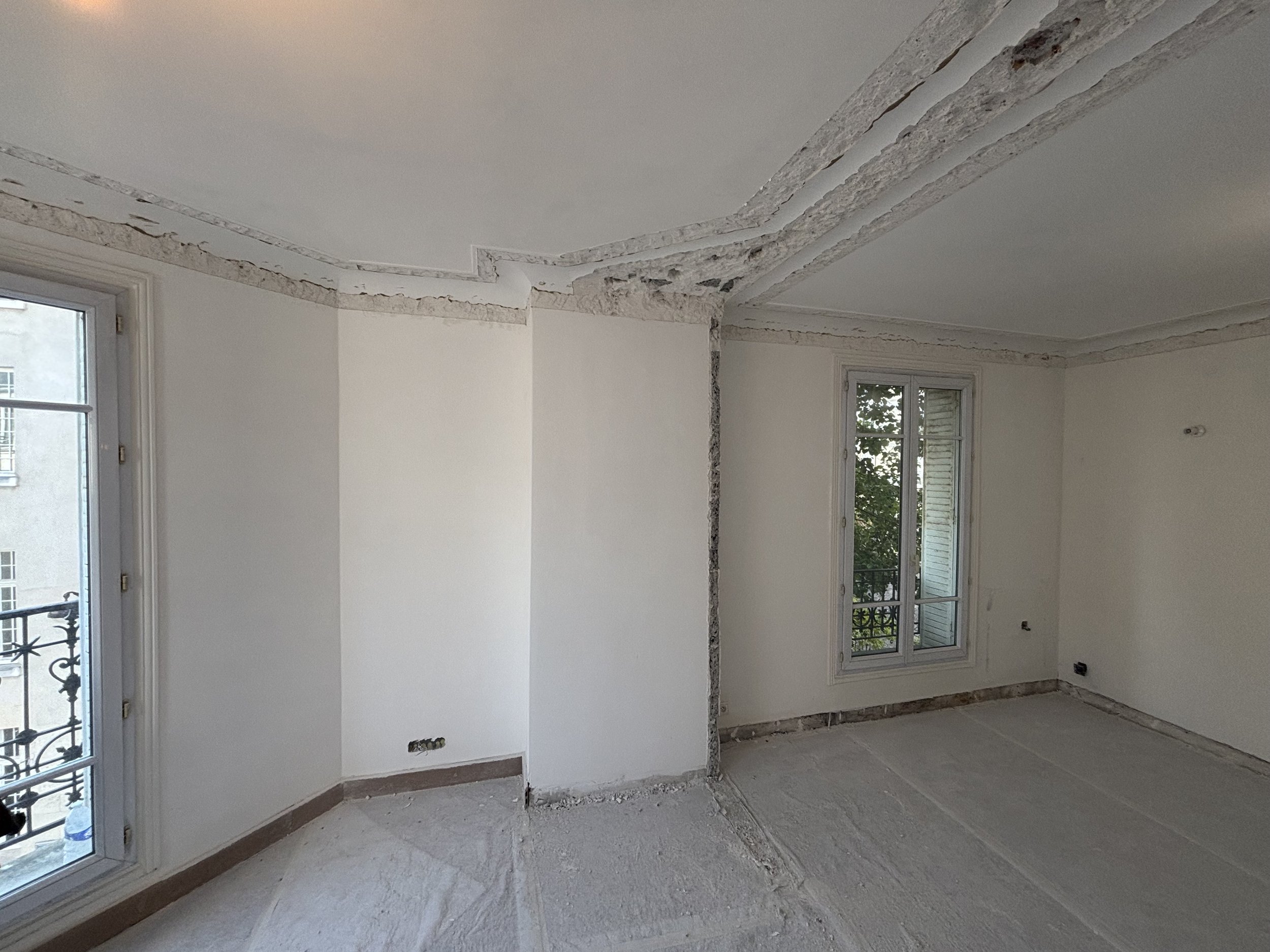 Une pièce en cours de rénovation avec des murs peints en blanc, deux fenêtres, et des travaux de plafonnage visibles sur le plafond et les murs, sans revêtement de sol installé.