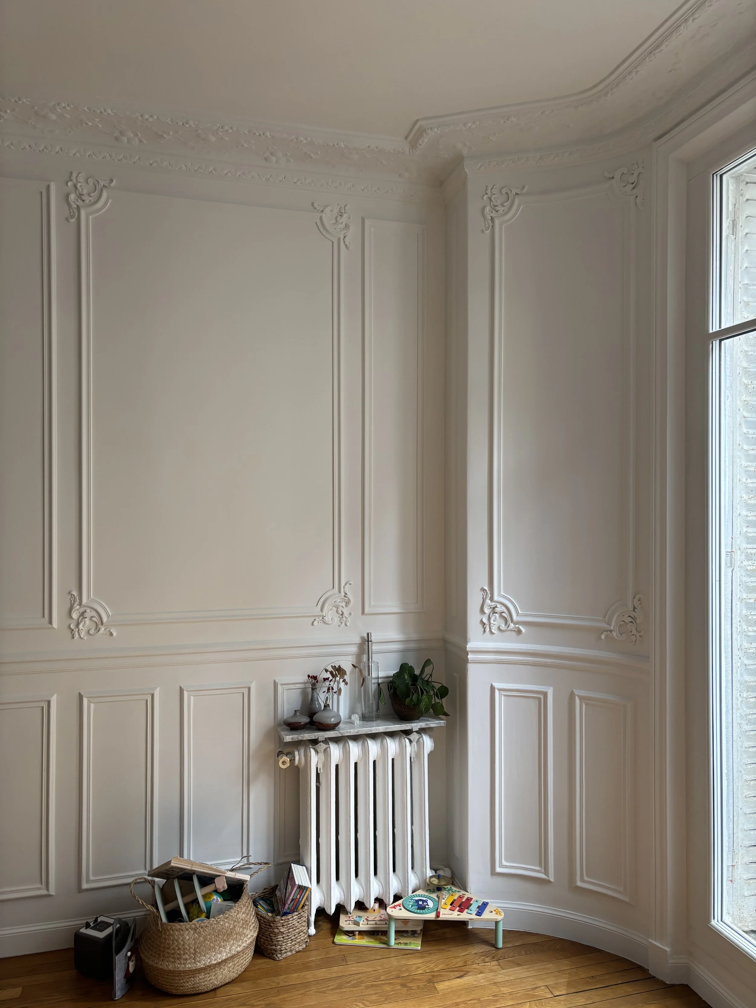 Coin d'une pièce avec murs blancs ornés de moulures, sol en bois, radiateur blanc, plantes et jouets pour enfants