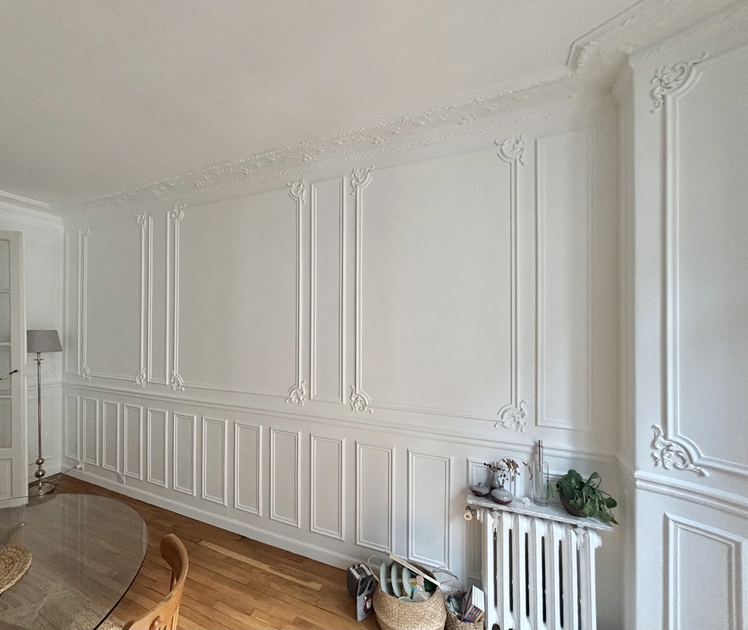 Salle à manger élégante avec moulures décoratives blanches sur les murs et plafond, sol en bois clair, espace organisé avec quelques objets décoratifs et une plante sur un radiateur.