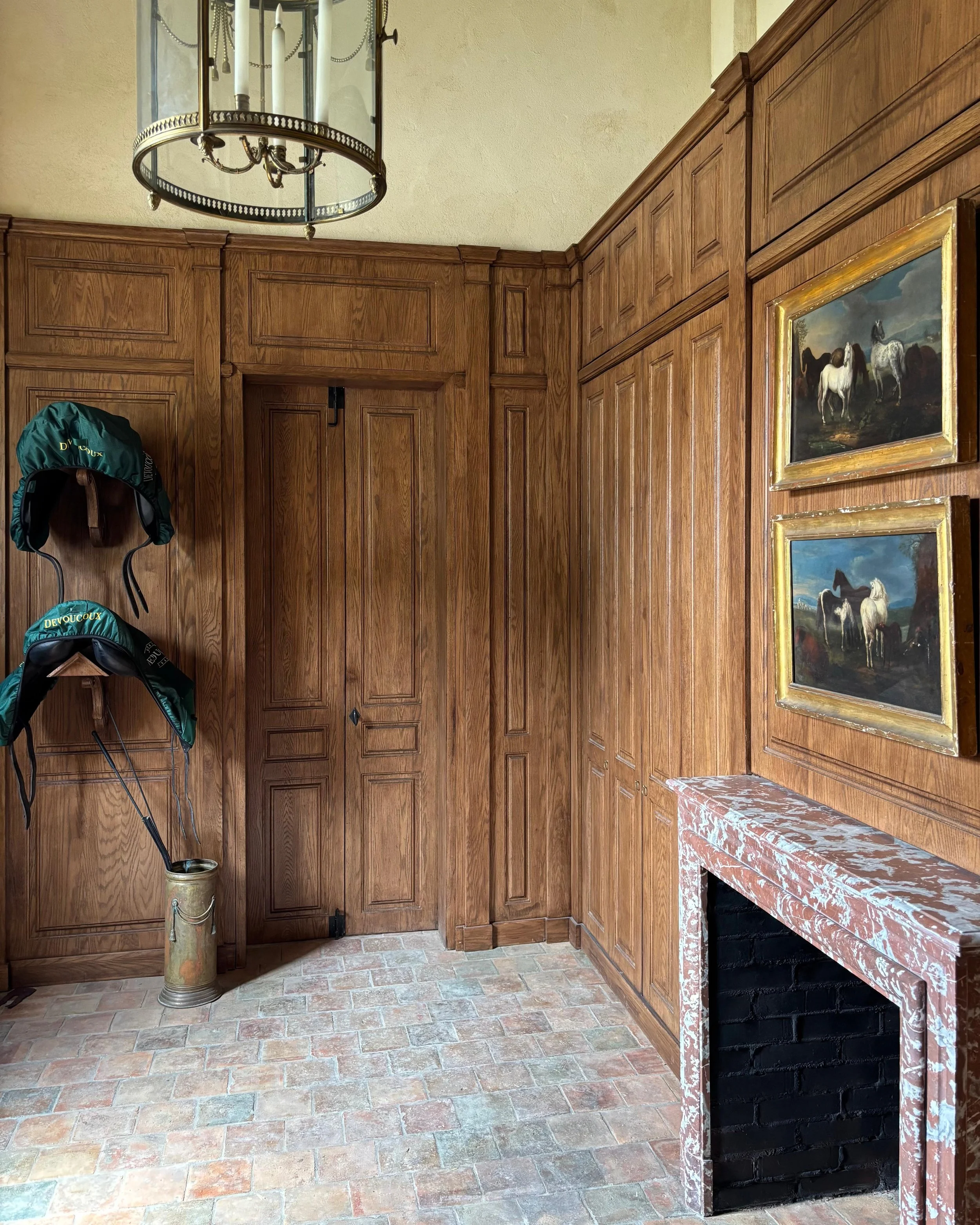 Une sellerie avec un mur recouvert de bois, une cheminée en marbre rose et deux tableaux de chevaux encadrés. Deux casquettes vertes sont suspendues à un porte-manteau en bois. Le sol est en carreaux de pierre beige.