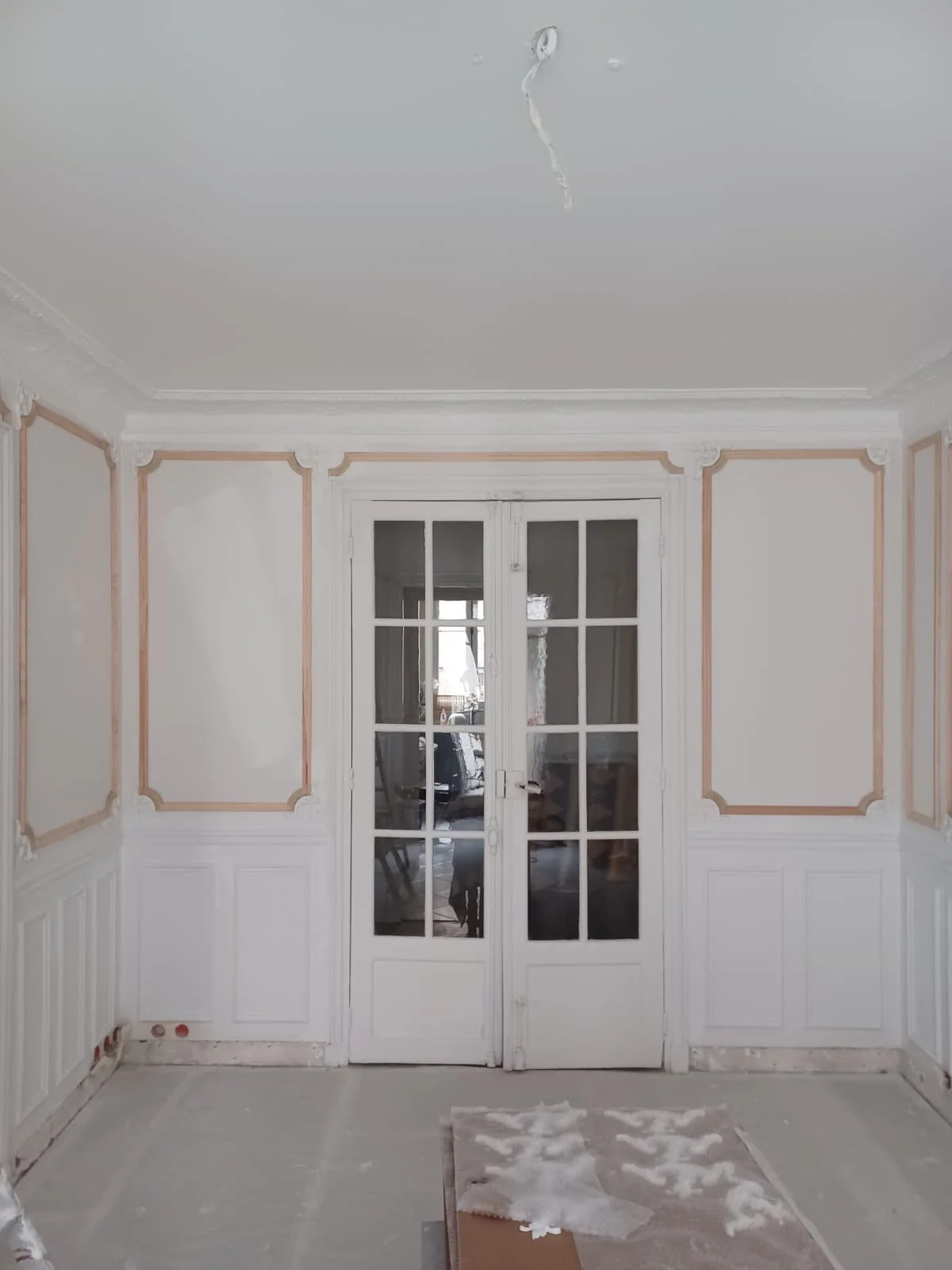 Une pièce avec des murs blancs, des portes-fenêtres en bois, des murs avec des moulures décoratives, et un sol en cours de rénovation avec des débris et un tapis.