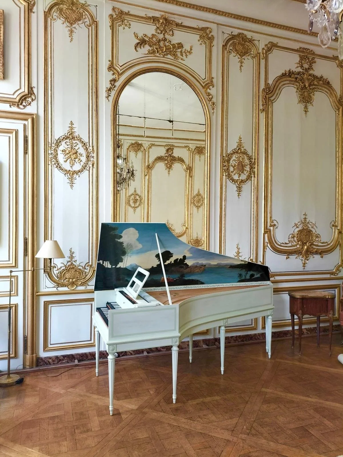 Un clavecin blanc avec une peinture représentant un paysage avec des arbres et une mer, placé dans une pièce élégante avec des murs ornementés en or et un parquet en bois.