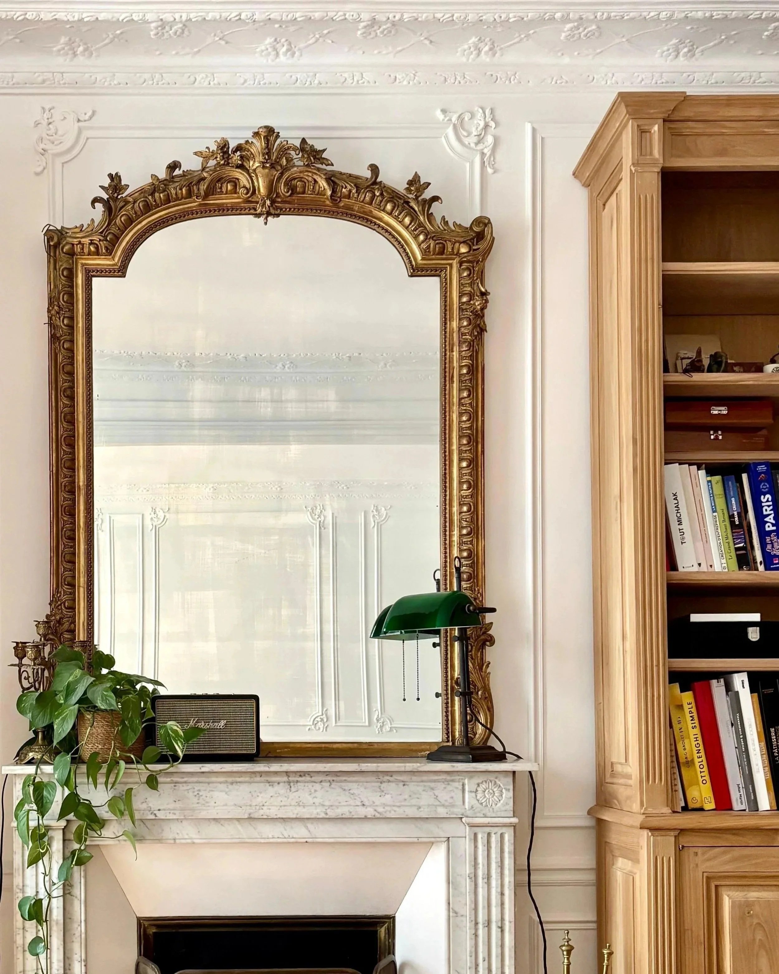 Grand miroir ancien avec cadre doré au-dessus d'une cheminée en marbre blanc, décorée de livres et d'une lampe verte de bureau, à côté d'une bibliothèque en bois.