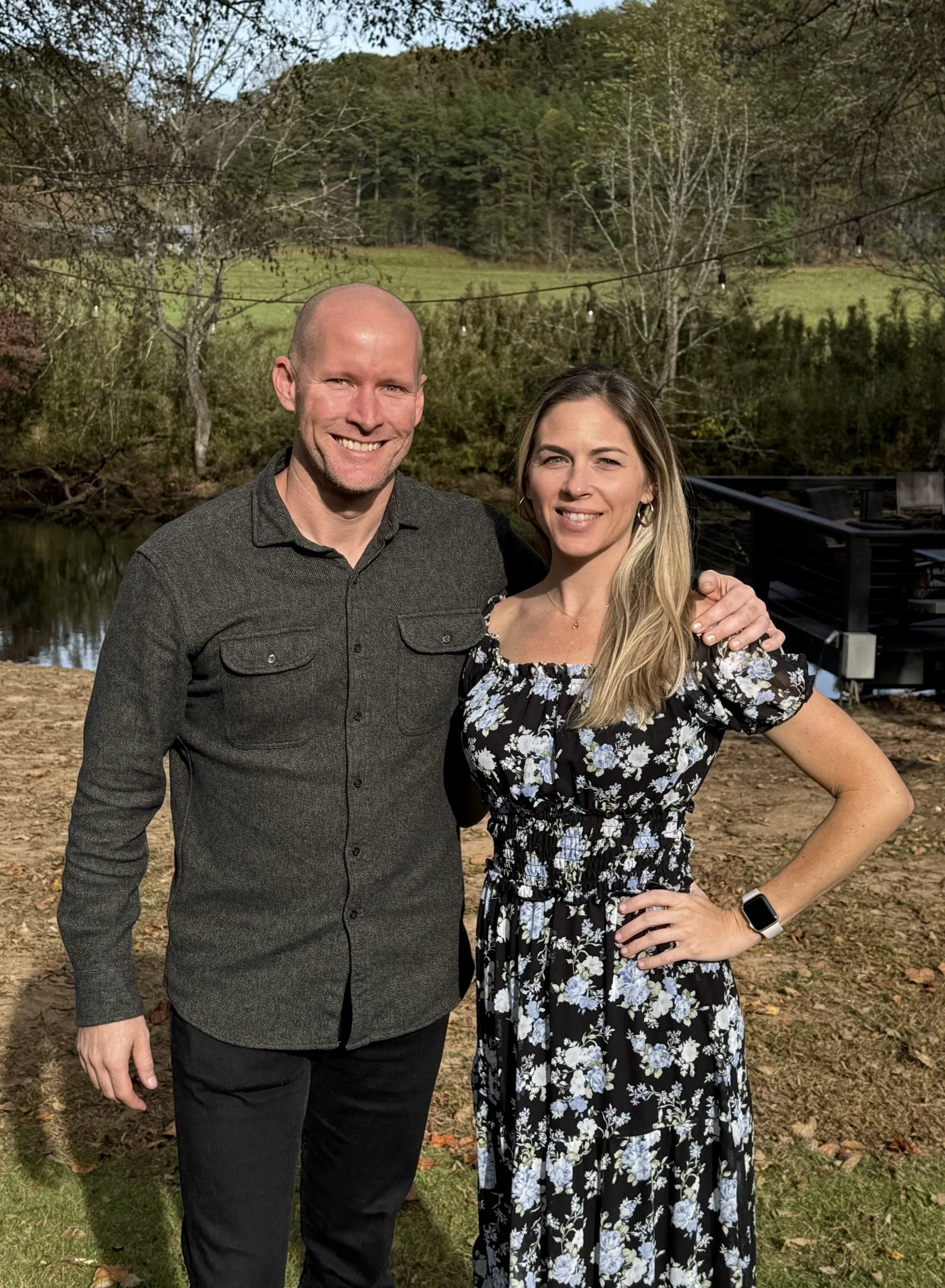 A smiling man and woman stand outdoors near a pond, with trees and a hillside in the background. The man has a shaved head and is wearing a dark long-sleeve shirt, while the woman has long blonde hair and is wearing a black floral dress with an off-the-shoulder neckline. They are standing close together, with the man's arm around the woman's shoulder.