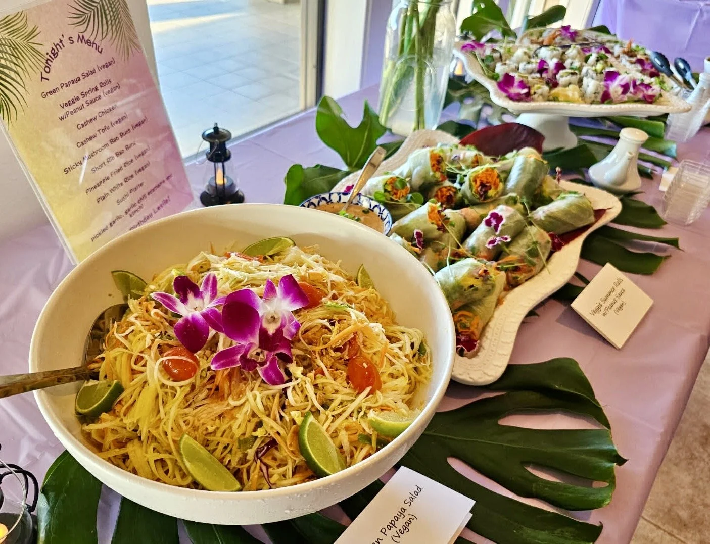 A buffet table with salads and spring rolls decorated with purple orchids and green leaves, with a menu and small signs labeling the dishes.