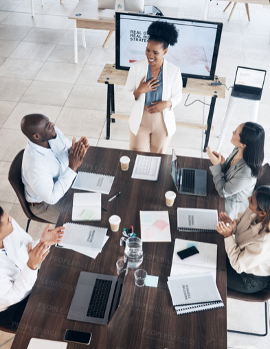 A woman stands in front of a group of five diverse professionals sitting around a conference table, giving a presentation. The table has laptops, notebooks, and cups of coffee.