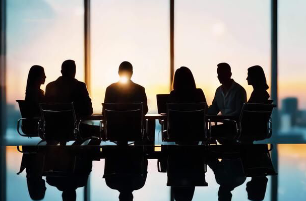 Silhouettes of six businesspeople sitting at a conference table in front of large window during sunset.