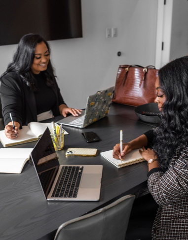 Two women sitting at a dark conference table with laptops, notebooks, and pens while working or having a discussion in a modern office.