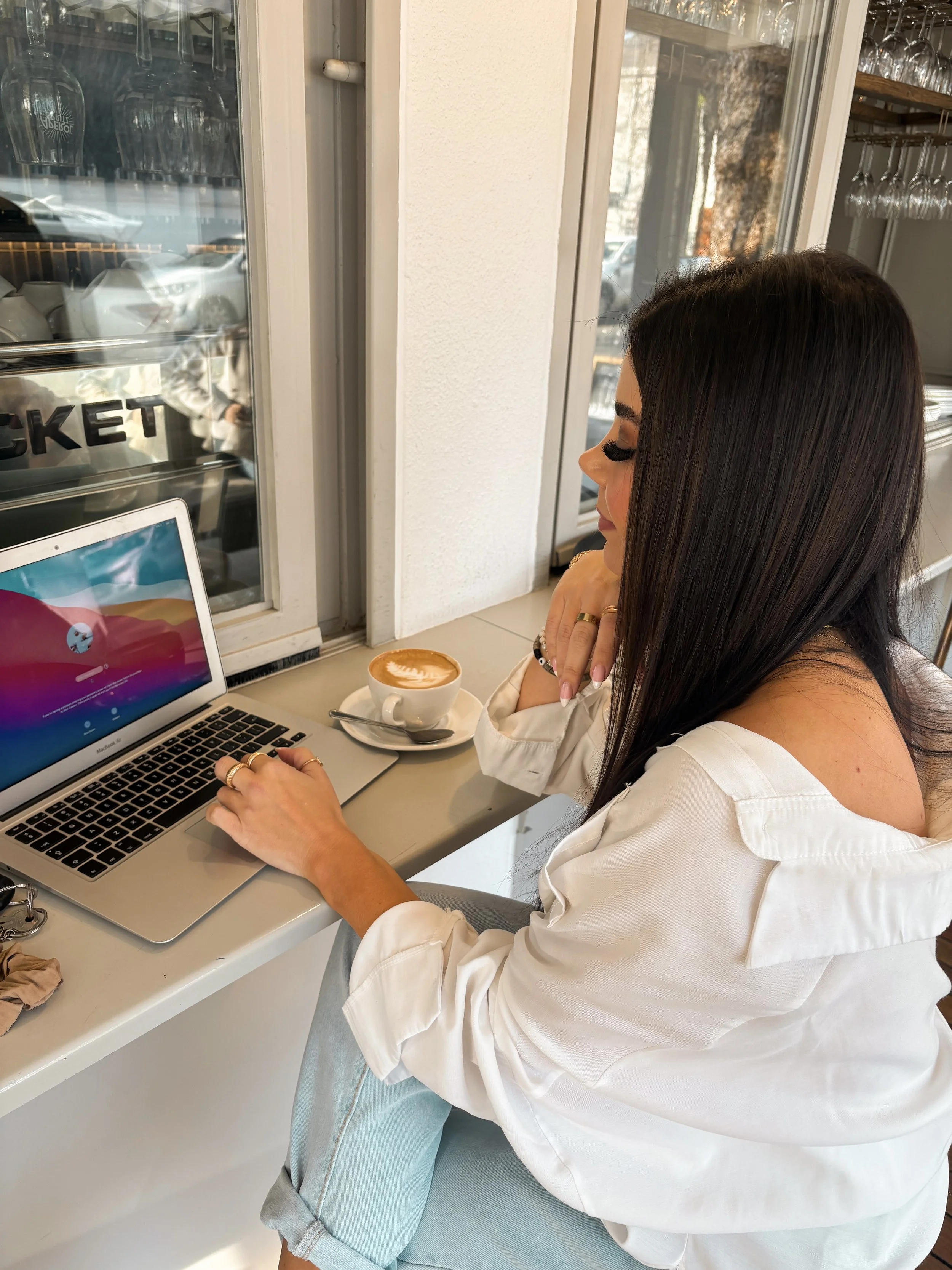A woman with long dark hair and a white shirt sitting at a cafe, looking at a laptop, with a coffee cup nearby, near a window.