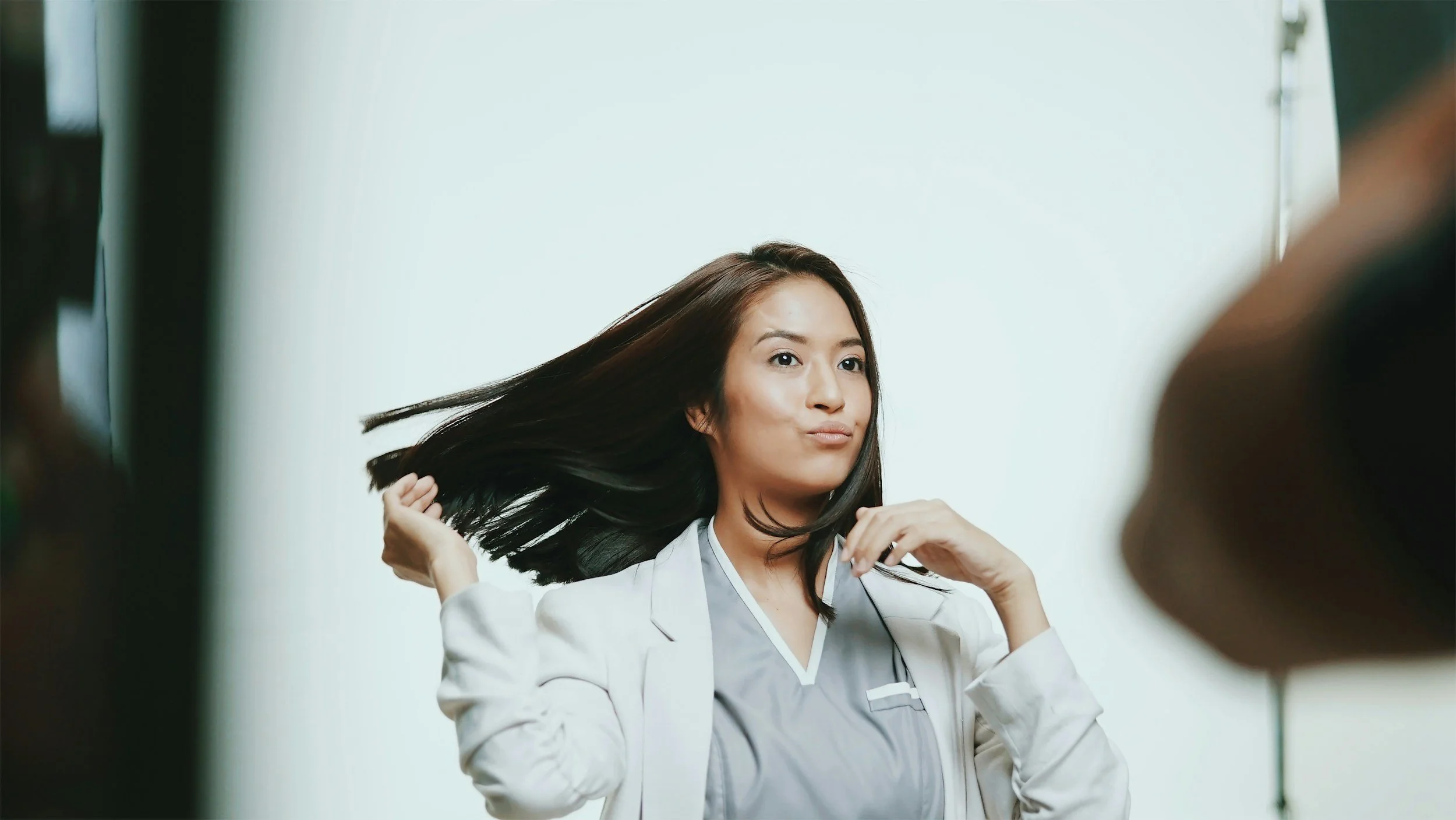 A woman with long, dark hair posing during a photo shoot, flipping her hair to the side in front of a plain light-colored background.