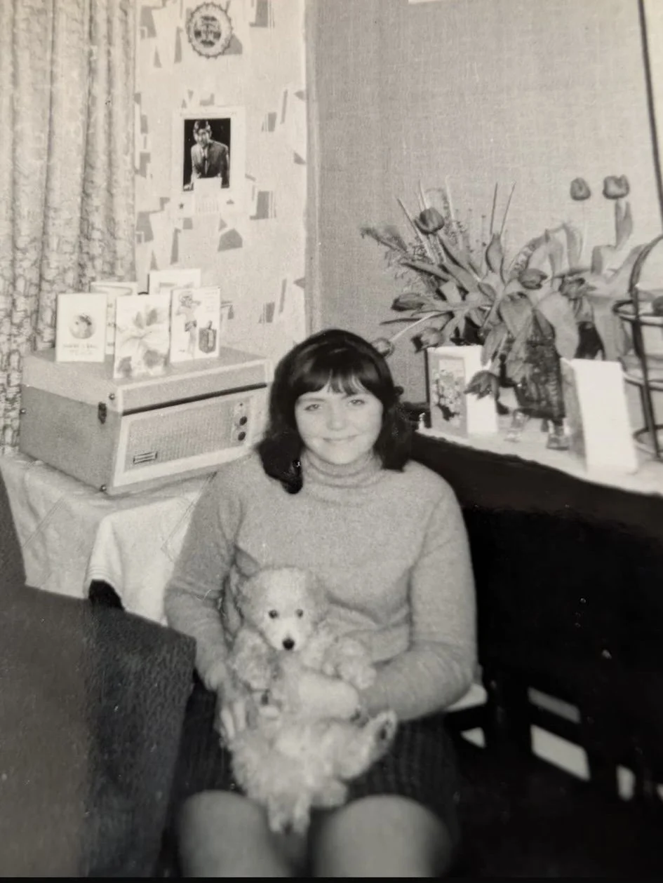 A woman with dark hair sitting and smiling, holding a small puppy. Behind her, a table with books, cards, and a large plant. On the wall, there's a photo of a man and some decorations.