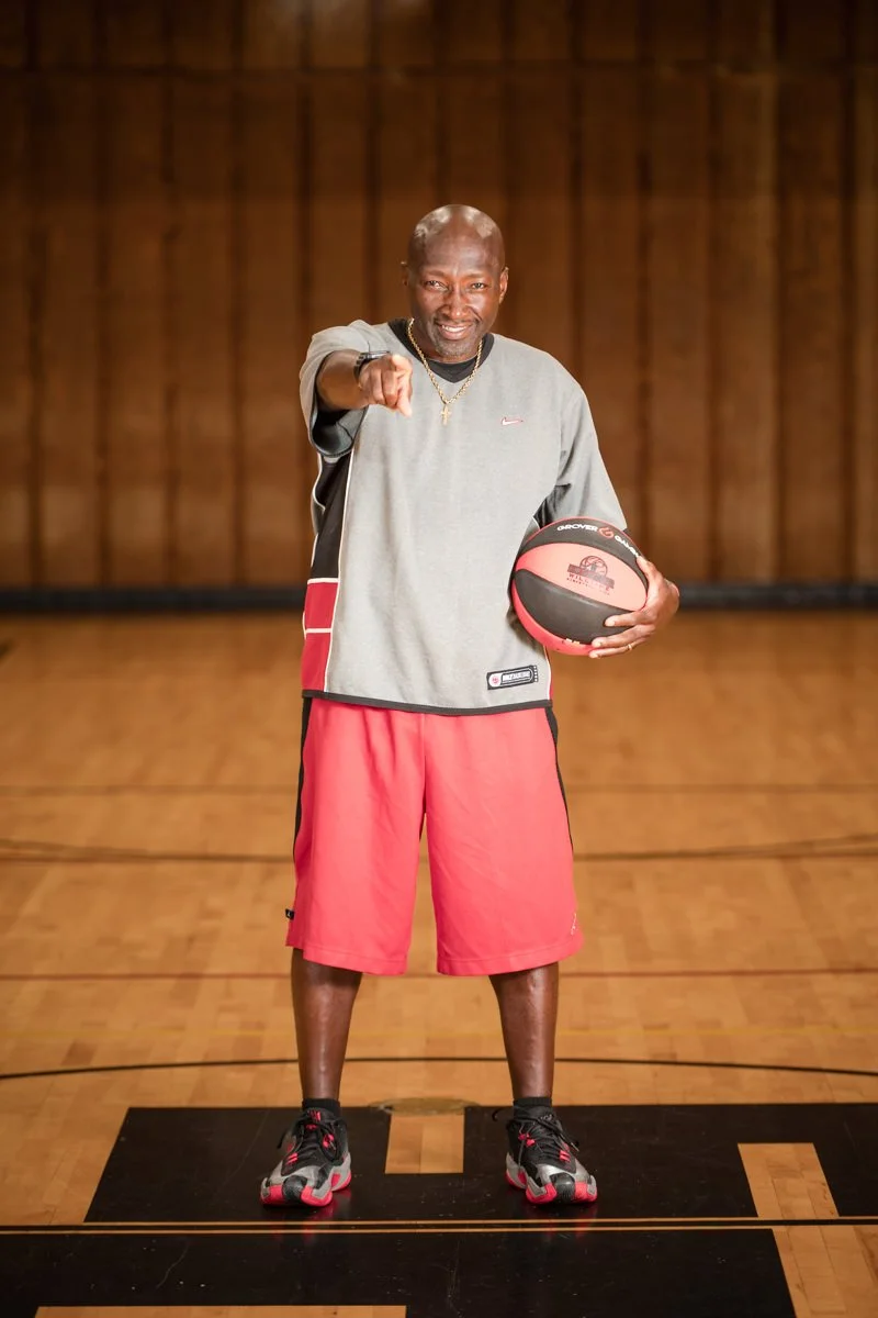 A man standing on a basketball court holding a basketball and pointing at the camera, wearing sportswear and basketball shoes.