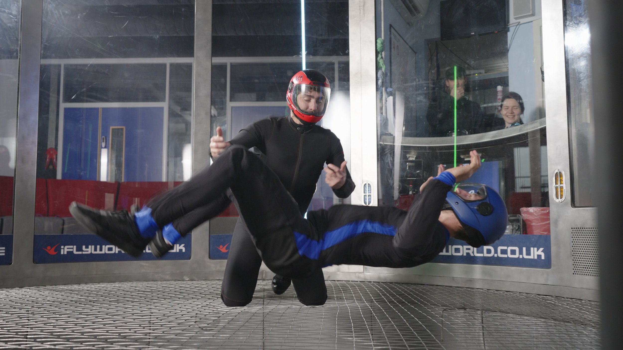 Two individuals are engaged in an indoor skydiving activity, with one person standing and the other person in a horizontal position, surrounded by a vertical wind tunnel.