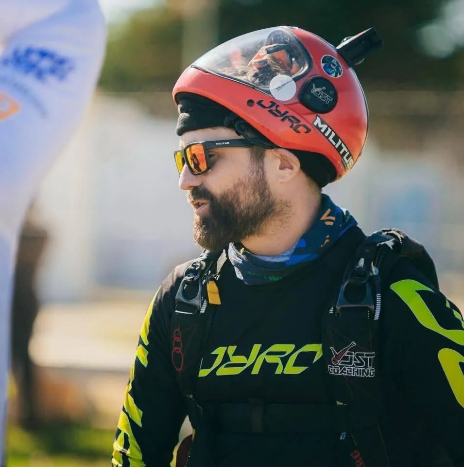 A man wearing a helmet, sunglasses, and a black and yellow motorcycle racing suit, with a backpack, sits outdoors on a sunny day.