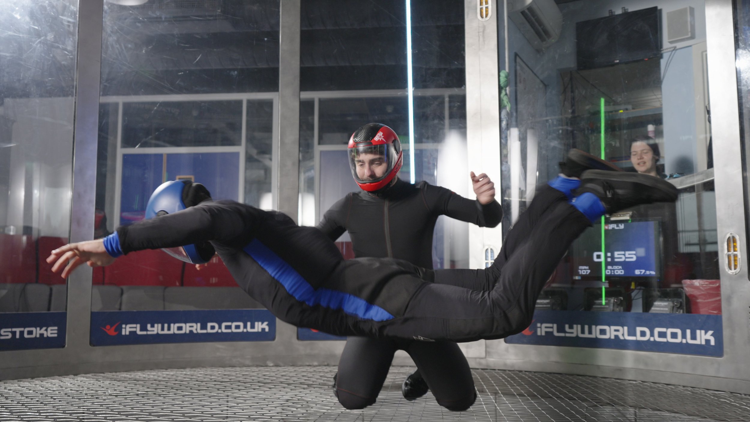 Two skydivers inside a vertical wind tunnel with one person supporting the other in a horizontal flying position. Both are wearing helmets, jumpsuits, and gloves. The scene is indoors with a glass wall and digital display showing speed and other data.