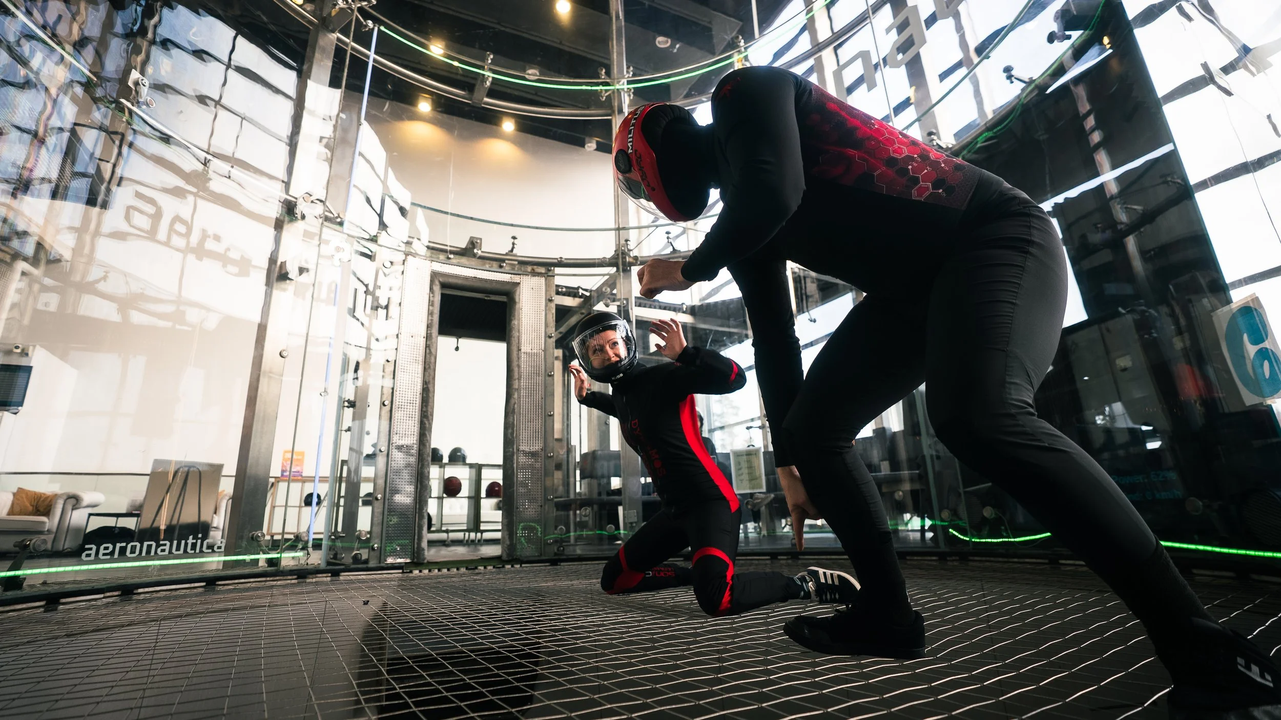 Two individuals in a skydiving simulator, wearing helmets and black suits with red accents, are inside a glass chamber with a netted floor, preparing for a skydiving experience.