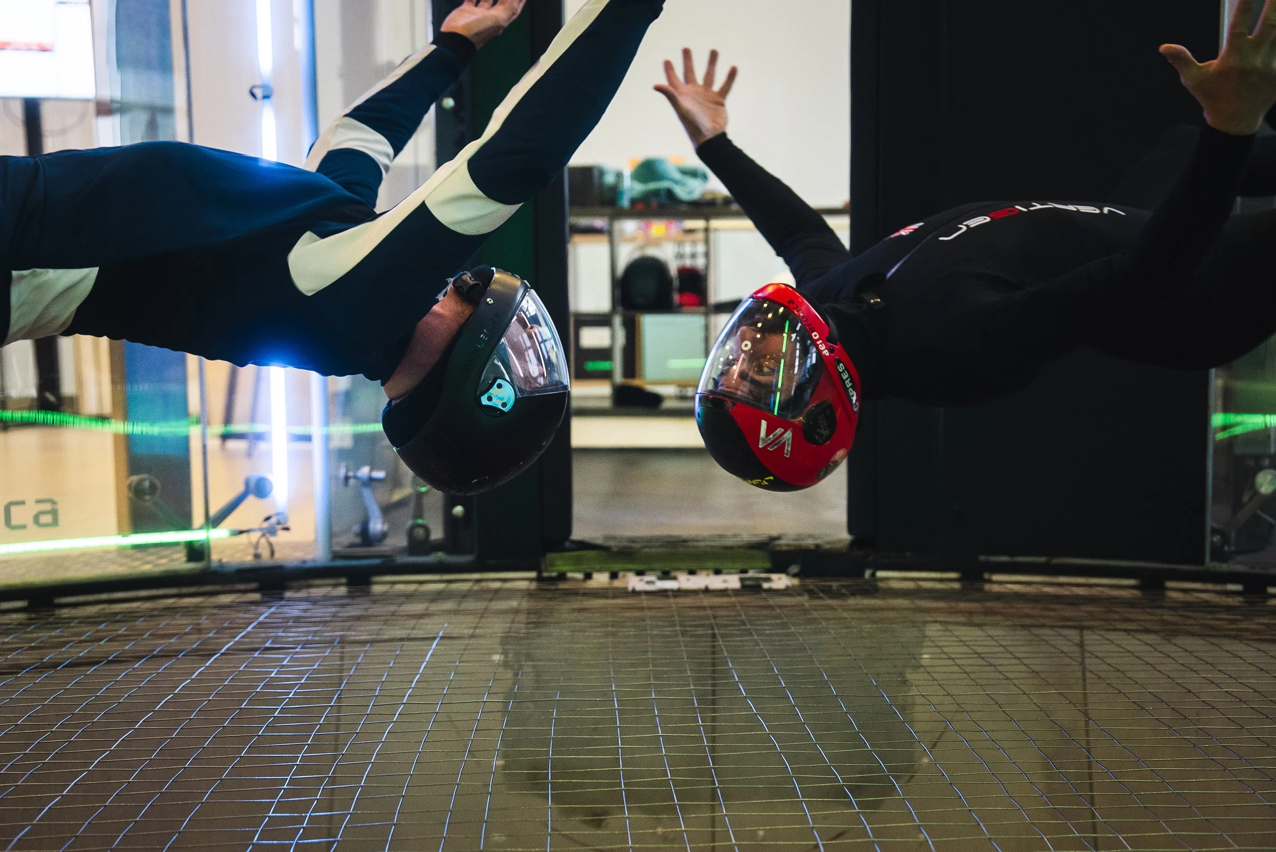 Two people in black racing helmets high-five each other in an indoor setting, with grid-like flooring and shelves in the background.