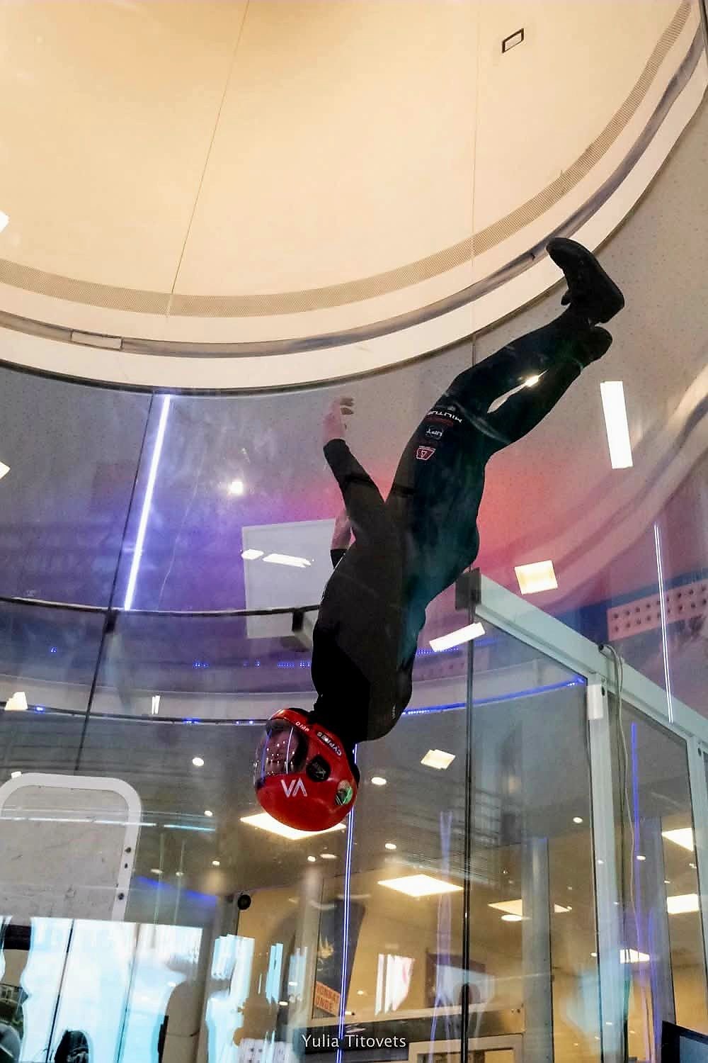 A person wearing a red helmet and black clothes upside down in a glass tunnel with a ceiling and floor visible.