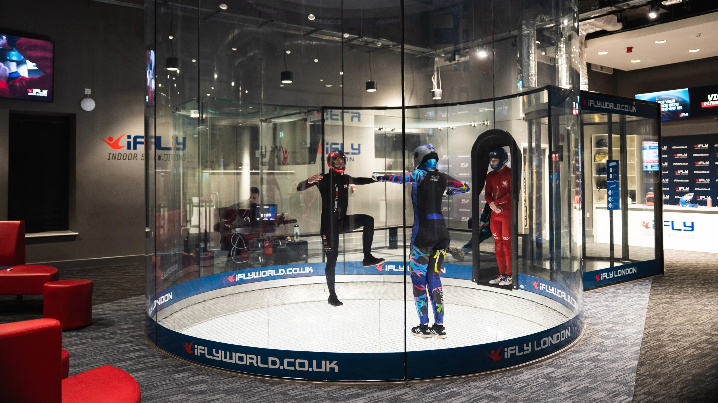 Indoor skydiving simulator with four people in colorful jumpsuits, two of whom are inside the glass tunnel and two outside, in a room with red chairs and a dark carpet. The background shows the iFLY London logo and screens.