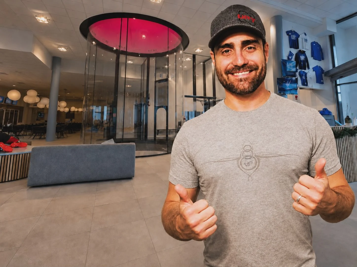 A smiling man wearing a gray t-shirt and a gray cap giving two thumbs up inside a modern indoor space with glass walls, hanging lights, and sports memorabilia on display.