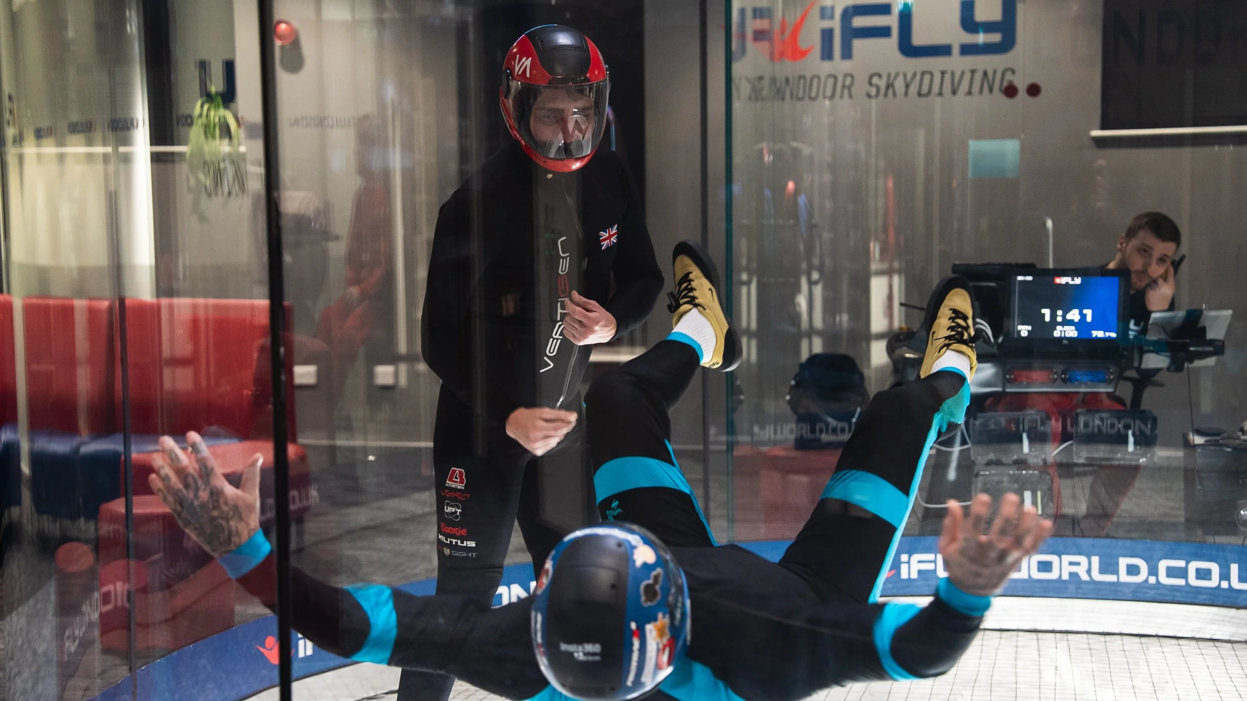 A person in a skydiving suit and helmet lying on the floor inside an indoor skydiving wind tunnel, with their legs raised and arms out, while another person in a black jacket and helmet is standing outside the tunnel looking inside and preparing to assist.