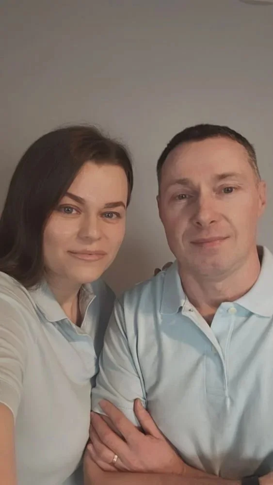A man and woman taking a selfie together against a plain wall, both wearing light-colored shirts.