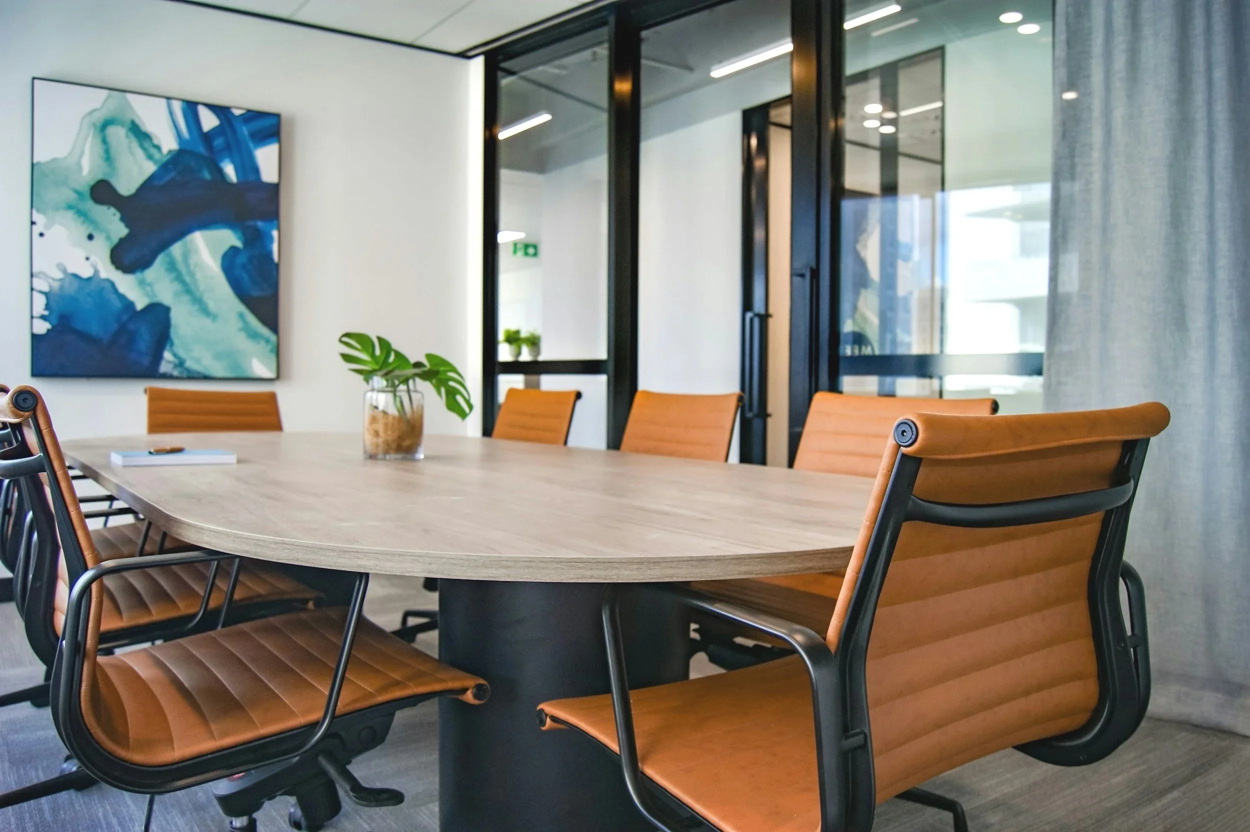 Modern conference room with a large wooden table, orange leather chairs, a plant centerpiece, abstract wall art, glass walls, and light gray curtains.
