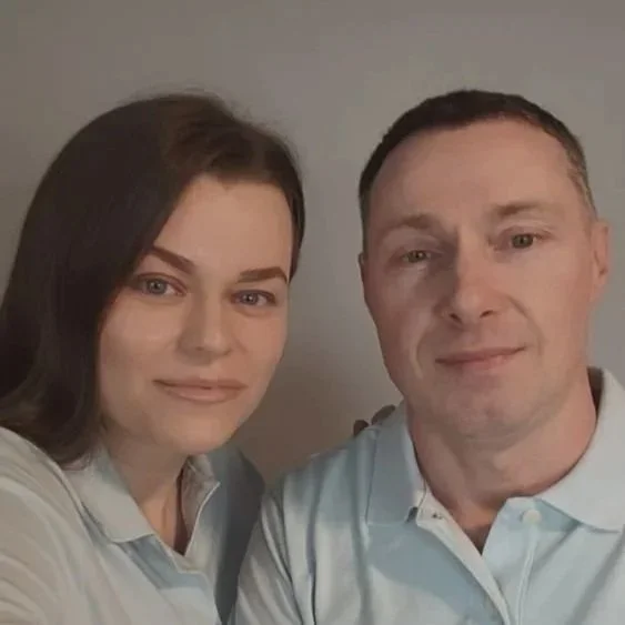 A man and woman taking a selfie together, both wearing light-colored collared shirts, smiling at the camera, with a plain background.