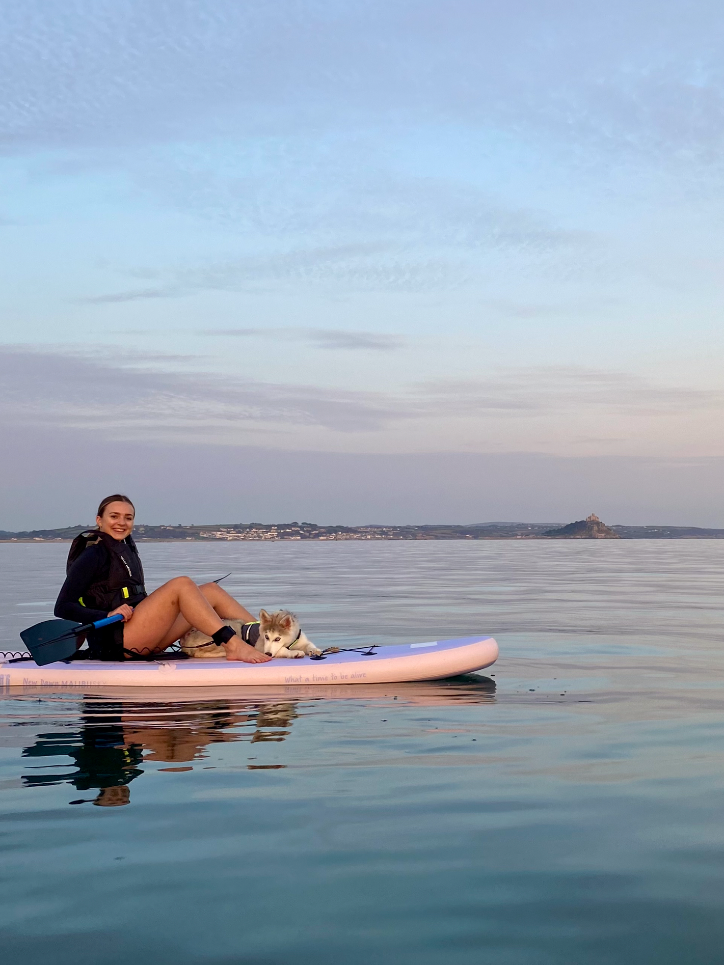 A young woman sitting on a paddleboard with a dog and paddle, smiling at the camera on a calm body of water during dusk, with a distant shoreline and a castle or fortress on a hill in the background.