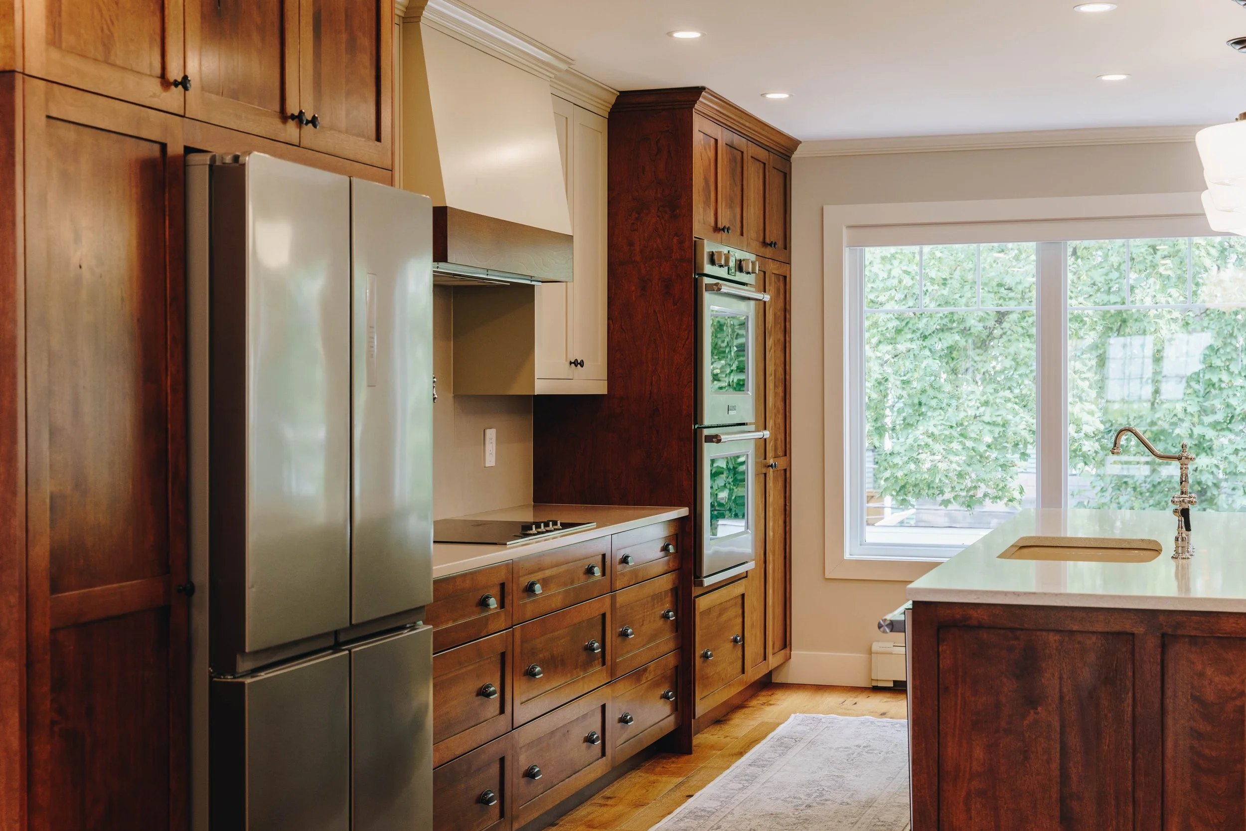 Modern kitchen with wooden cabinetry, stainless steel refrigerator, double oven, and large window overlooking trees.