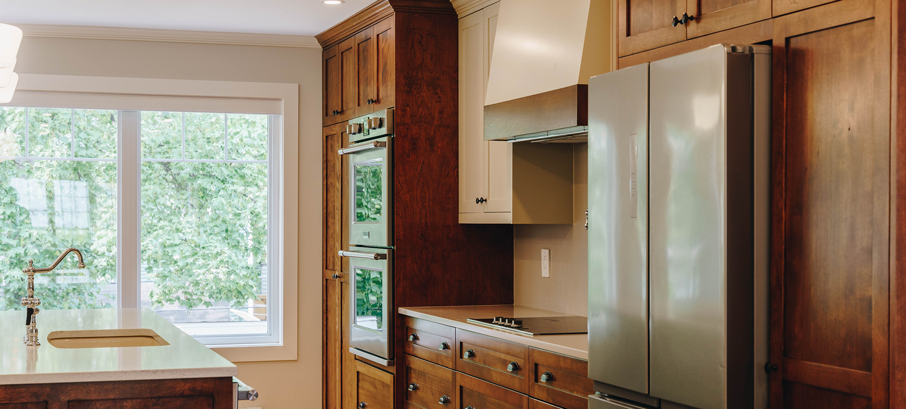 Kitchen with wooden cabinets, a stainless steel double oven, a beige countertop with a sink, a stove with an oven hood, and a large window showing green trees outside.