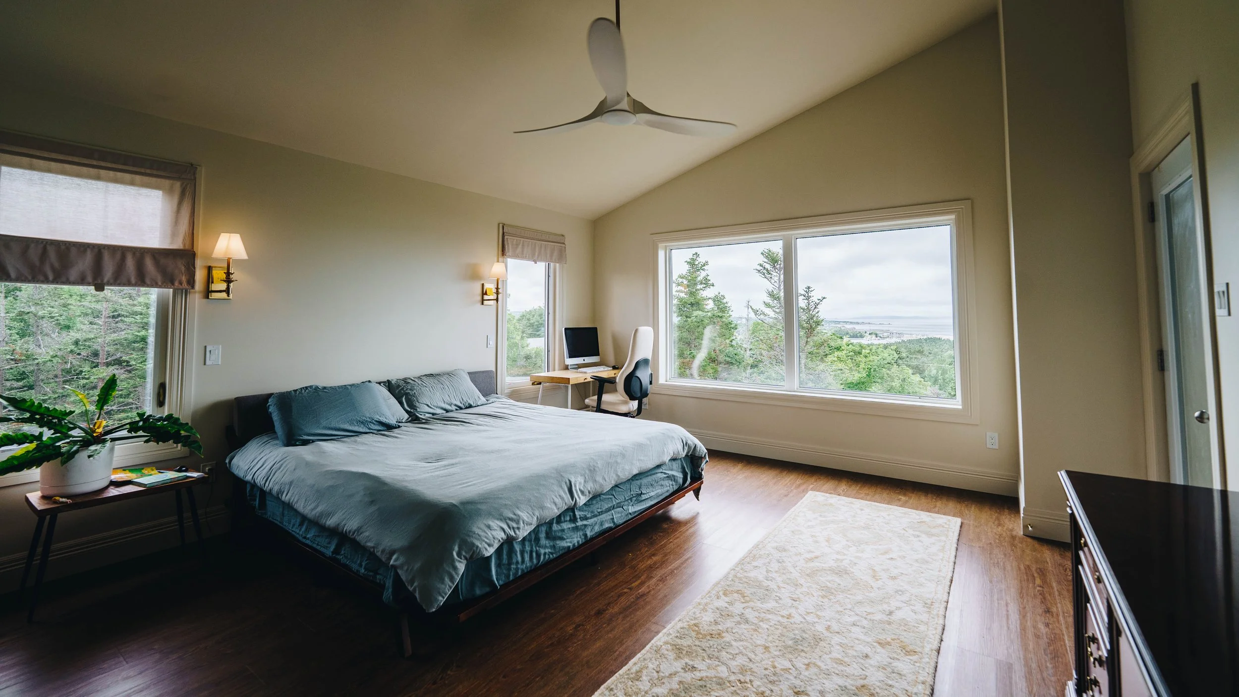 A bedroom with a large bed, nightstands, and a plant in a white pot. There are two wall-mounted lamps, a desk with a computer and a white office chair by a big window with a scenic view of trees and the outdoors. The room has hardwood flooring, a cei