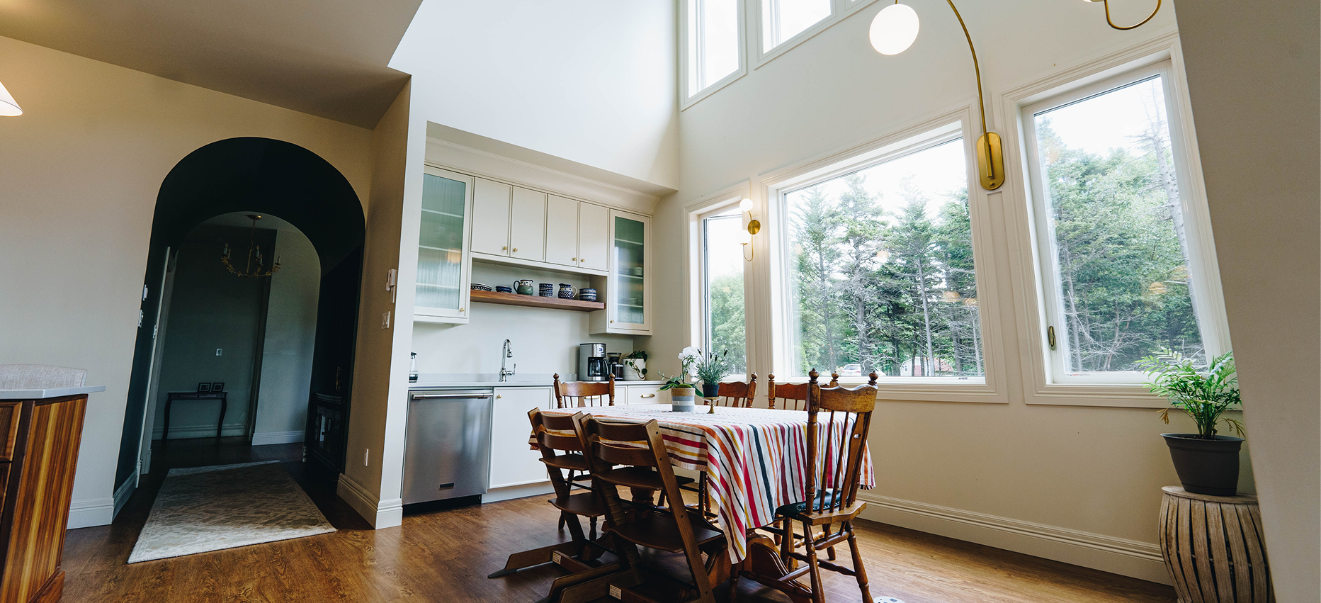 Bright kitchen dining area with a wooden table covered with a striped tablecloth, surrounded by wooden chairs, large windows letting in natural light, potted plants, white cabinets, and wooden flooring.
