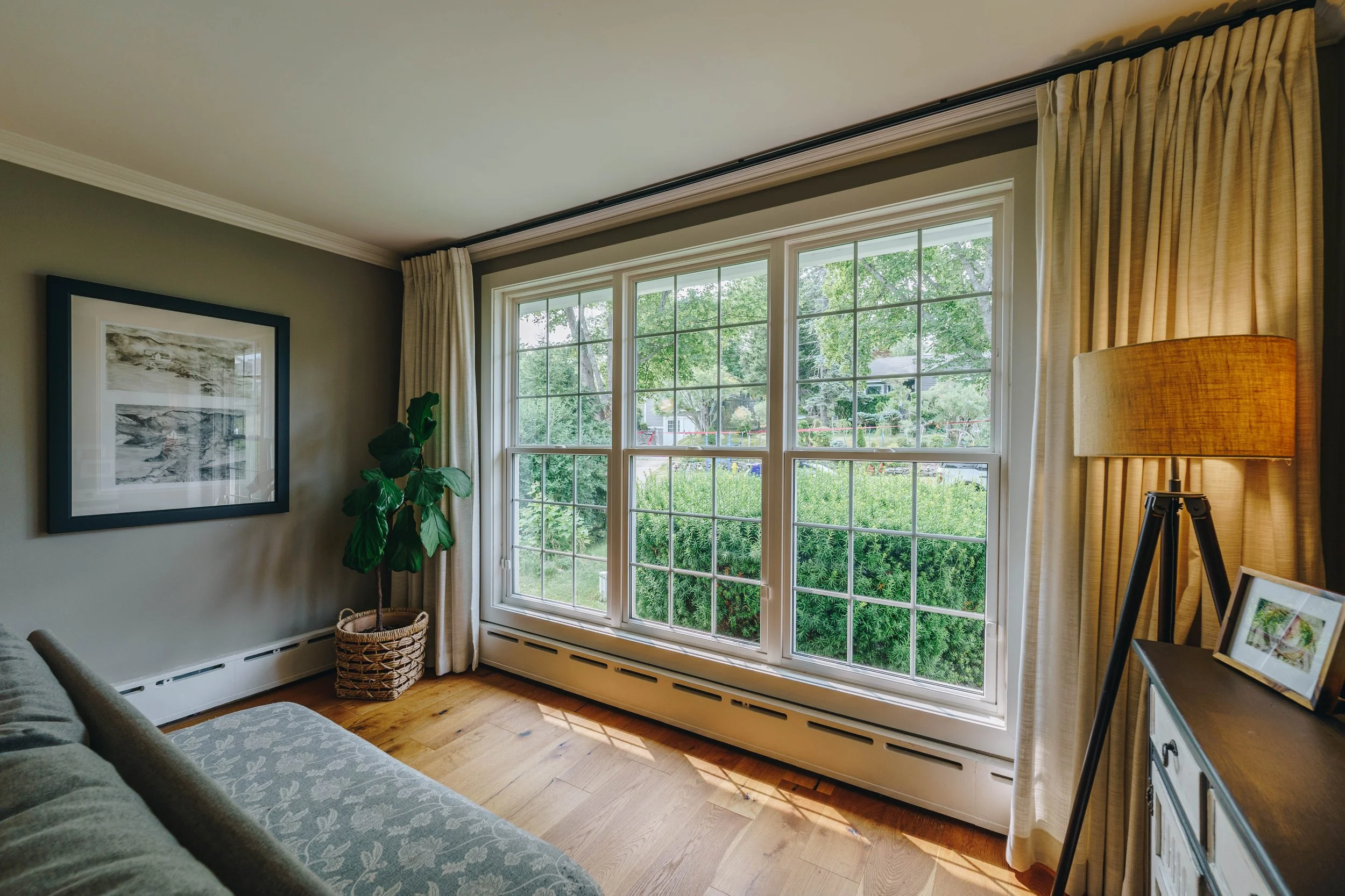 Living room with large window overlooking a garden, with a sofa on the left, framed artwork on the wall, a potted plant in a basket, and a lamp on a side table.
