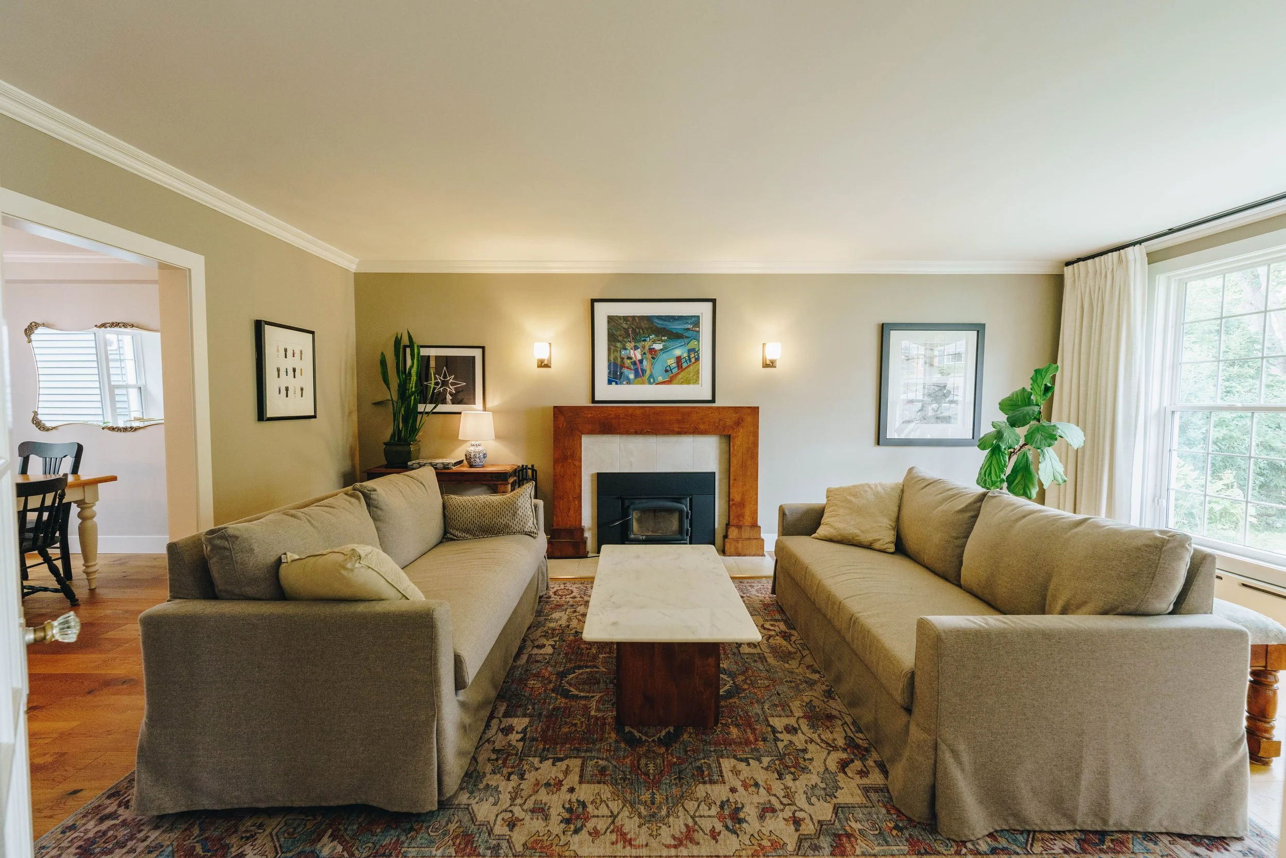 Living room with two beige sofas facing each other, a central marble coffee table, a fireplace with a wooden mantel, wall art, a potted plant, and windows with curtains allowing natural light.