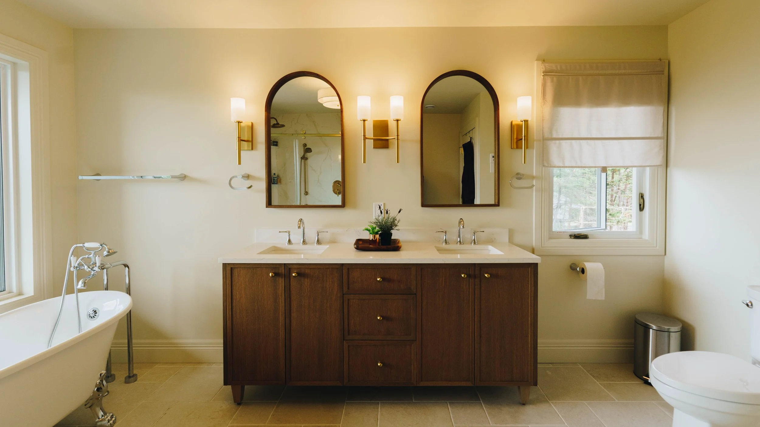 Bathroom with a double vanity, two mirrors, and wall-mounted lights, a bathtub on the left, and a window on the right