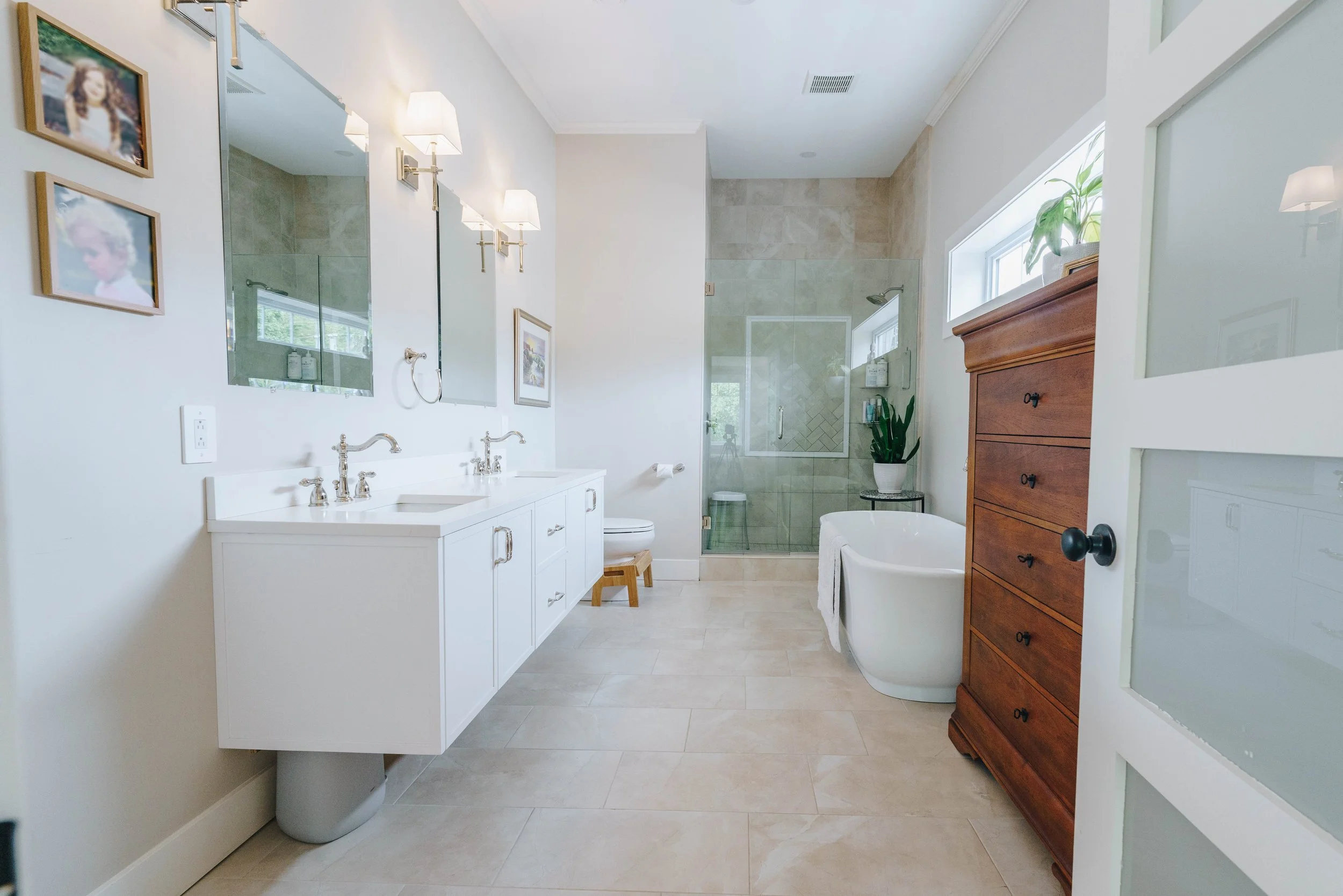 Bright bathroom with white double vanity, large mirror, wall-mounted lights, framed photos, toilet, glass-enclosed shower, standing bathtub, wooden dresser, potted plant, and window with natural light.
