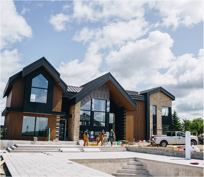 Modern house under construction with workers on site, large windows, a pool in the foreground, and a partly cloudy sky.