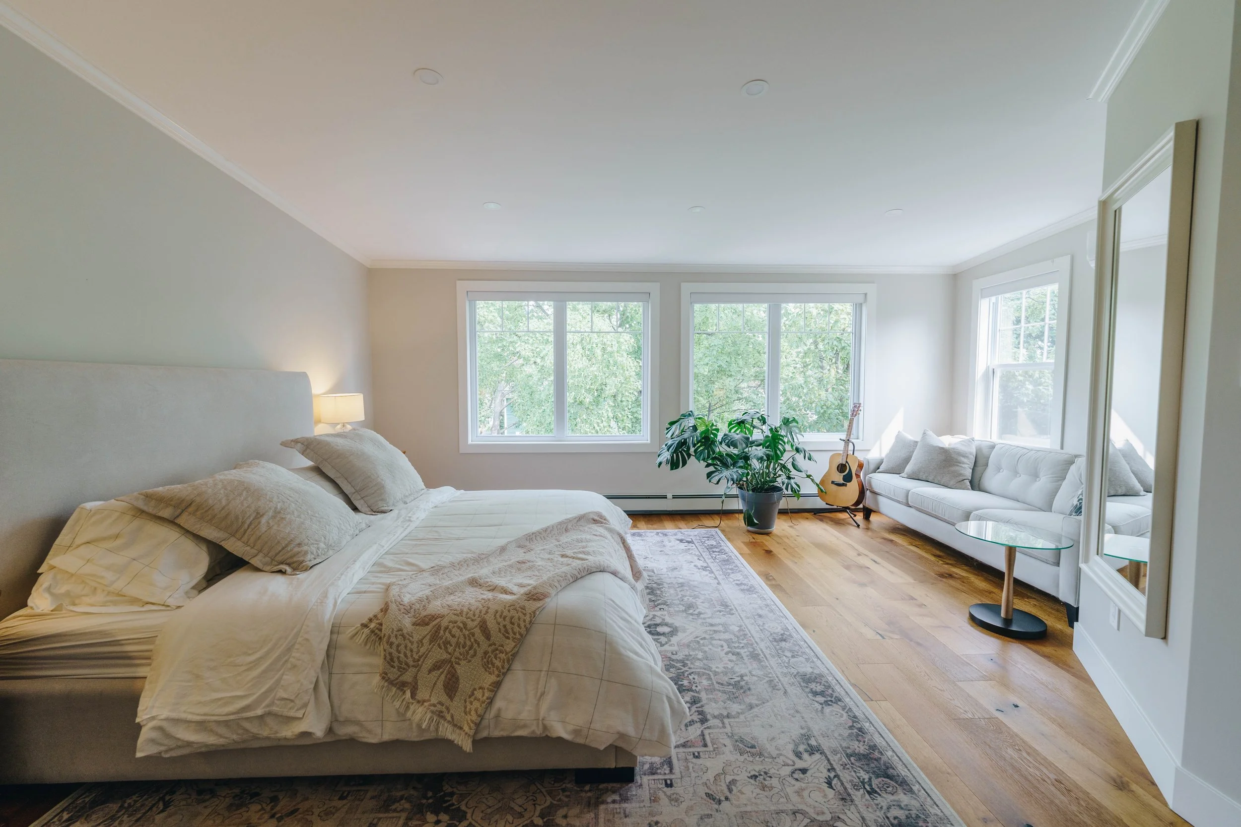 Bright bedroom with a large bed, light-colored bedding and pillows, a nightstand with a lamp, hardwood floors, a sitting area with a white sofa, a large mirror, a potted plant, an acoustic guitar, and large windows showing greenery outside.