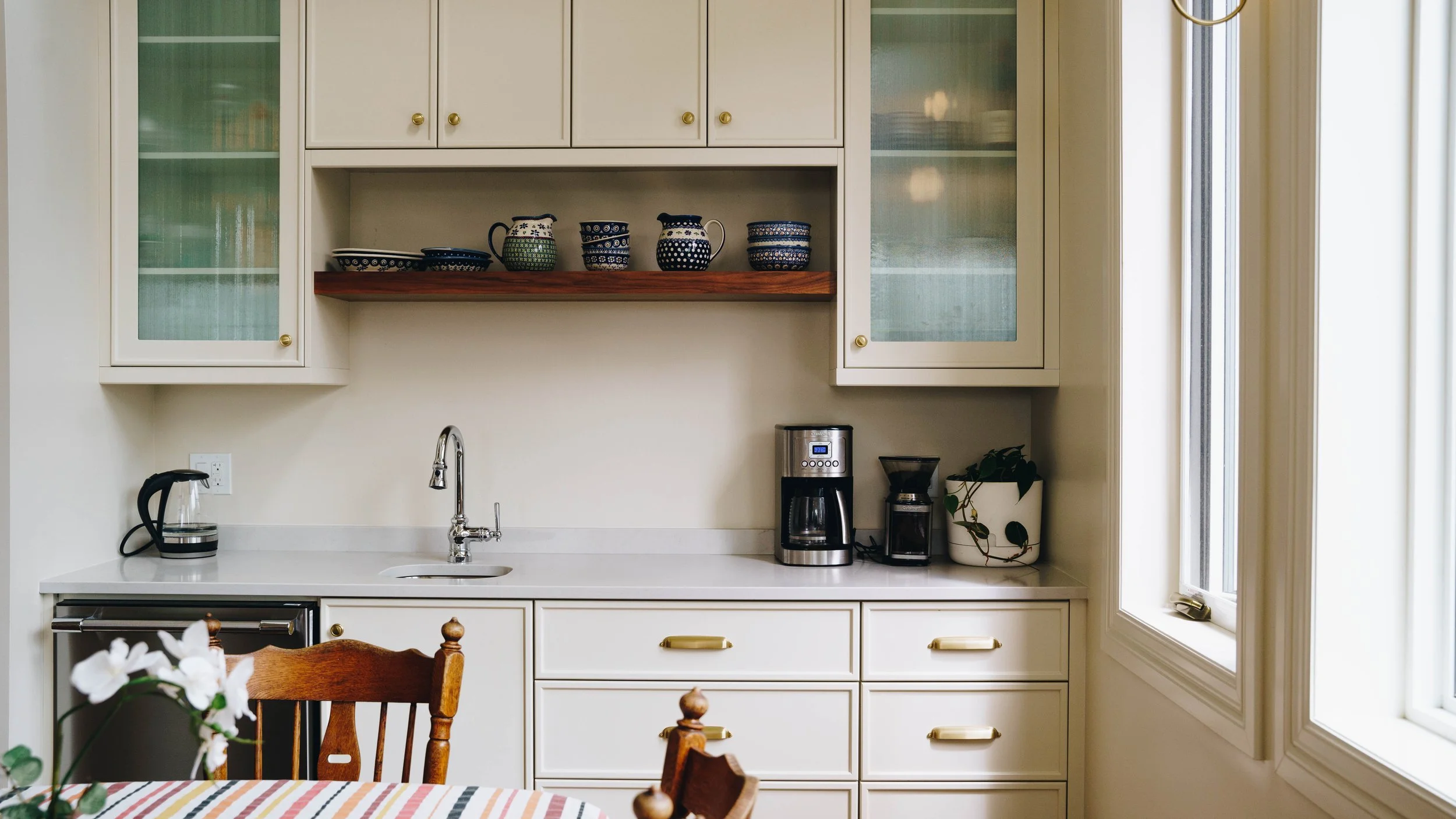 A kitchen with white cabinets, a countertop, coffee maker, coffee grinder, electric kettle, a potted plant, and a wooden chair in front of a window.