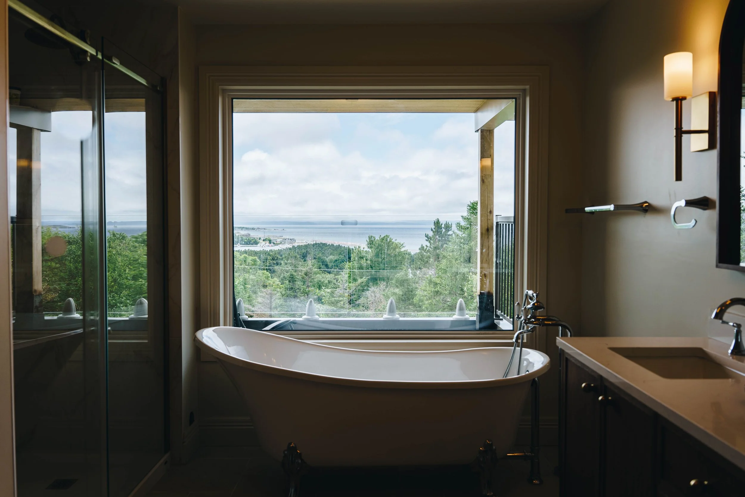Bathroom with a large window overlooking a lush green landscape and water in the distance, a white freestanding bathtub, a sink with a wooden cabinet, and modern wall-mounted lights.