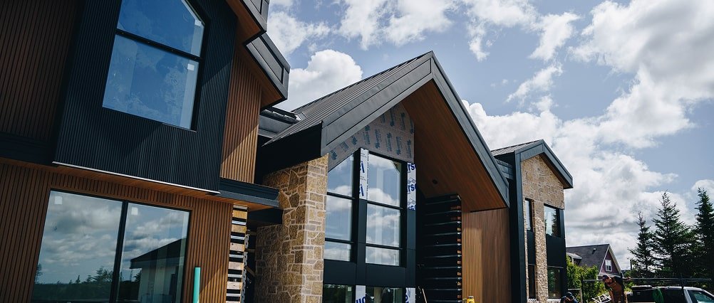 Modern residential building with large glass windows and a combination of wood and stone exterior, under a partly cloudy sky.