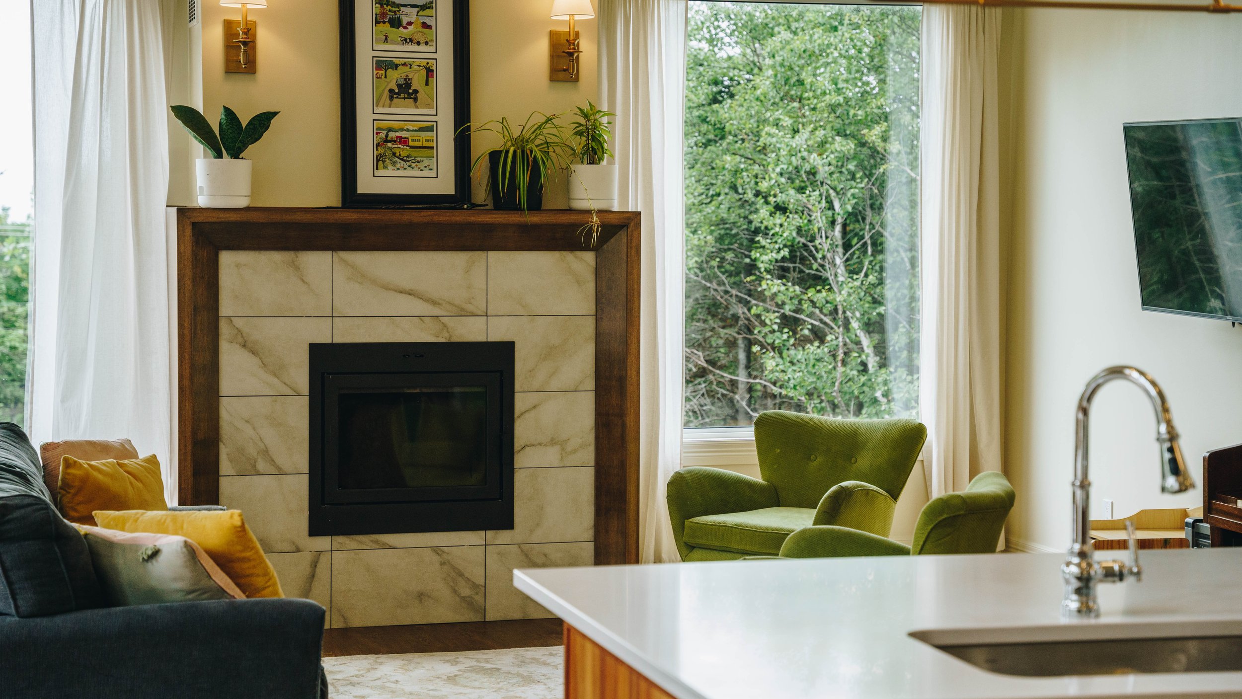 Living room with a fireplace, a television mounted on the wall, a window with trees outside, green armchairs, a dark sofa with yellow pillows, a white countertop with a sink and faucet, and decorative plants and framed art above the fireplace.